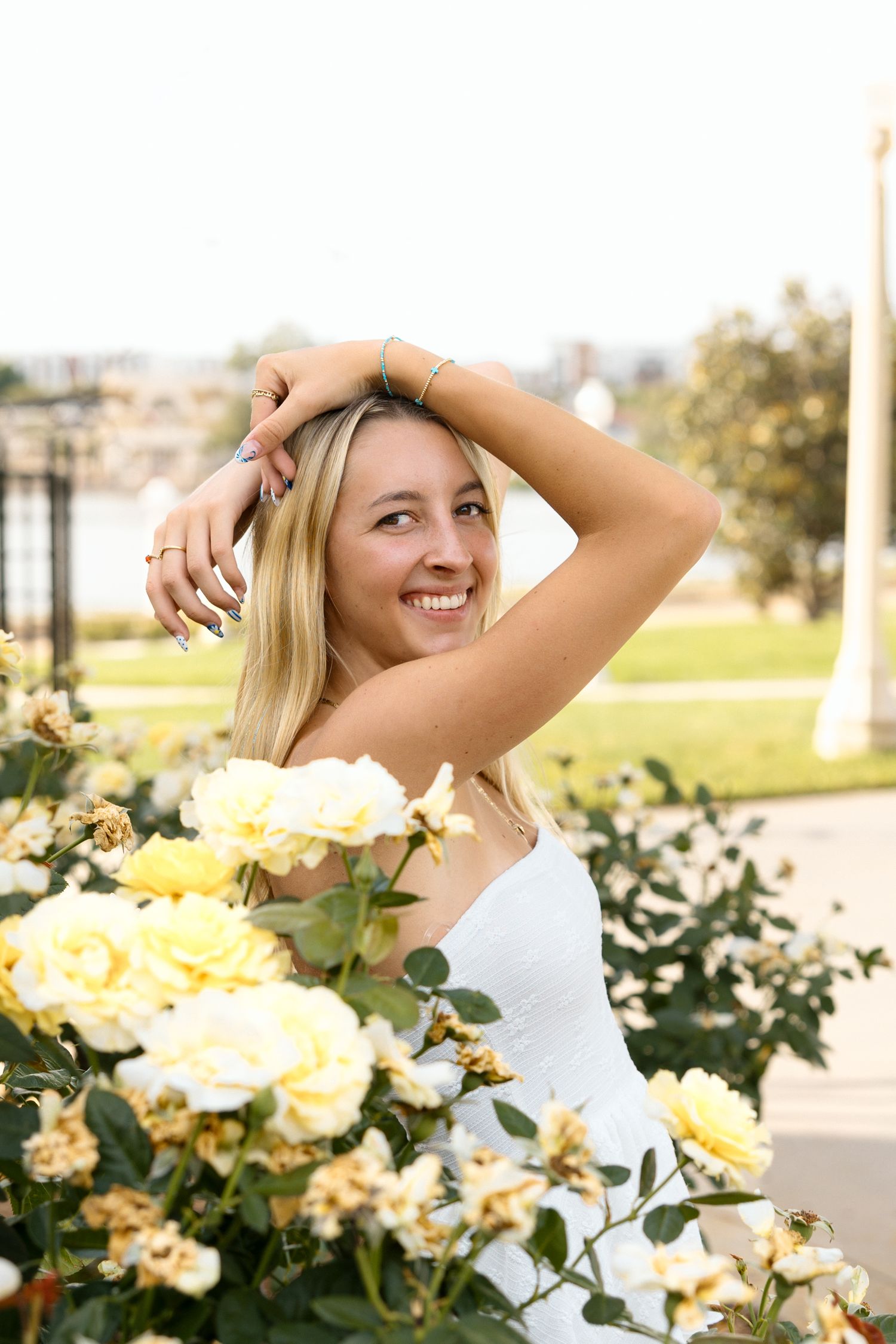 Smiling blonde woman in white dress posing among yellow roses in a sunny outdoor garden setting.