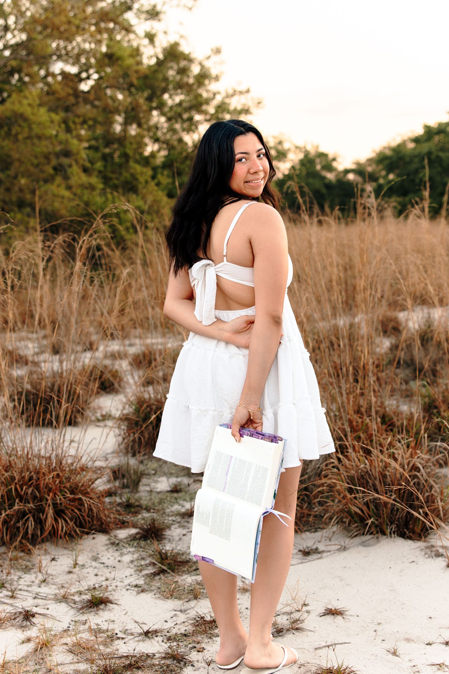 Young woman in white dress holding a book, standing on sandy beach with tall grass in golden hour light.