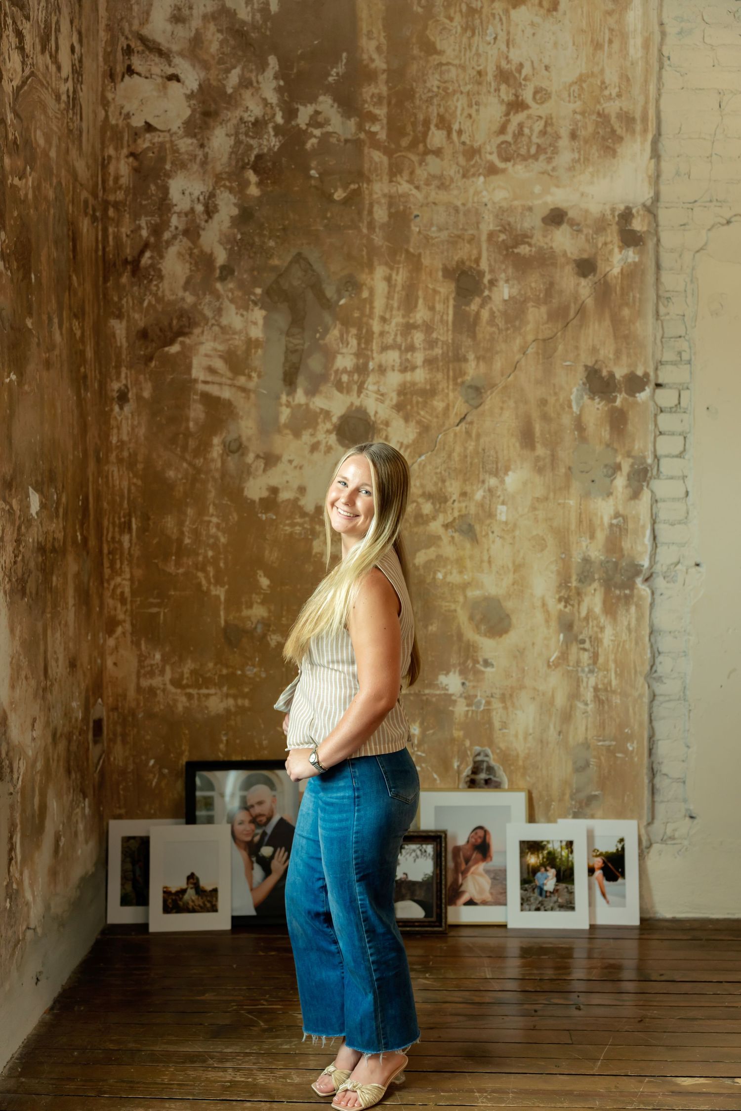 Pregnant woman in jeans and striped top smiling sideways, standing before rustic wall with framed family photos.