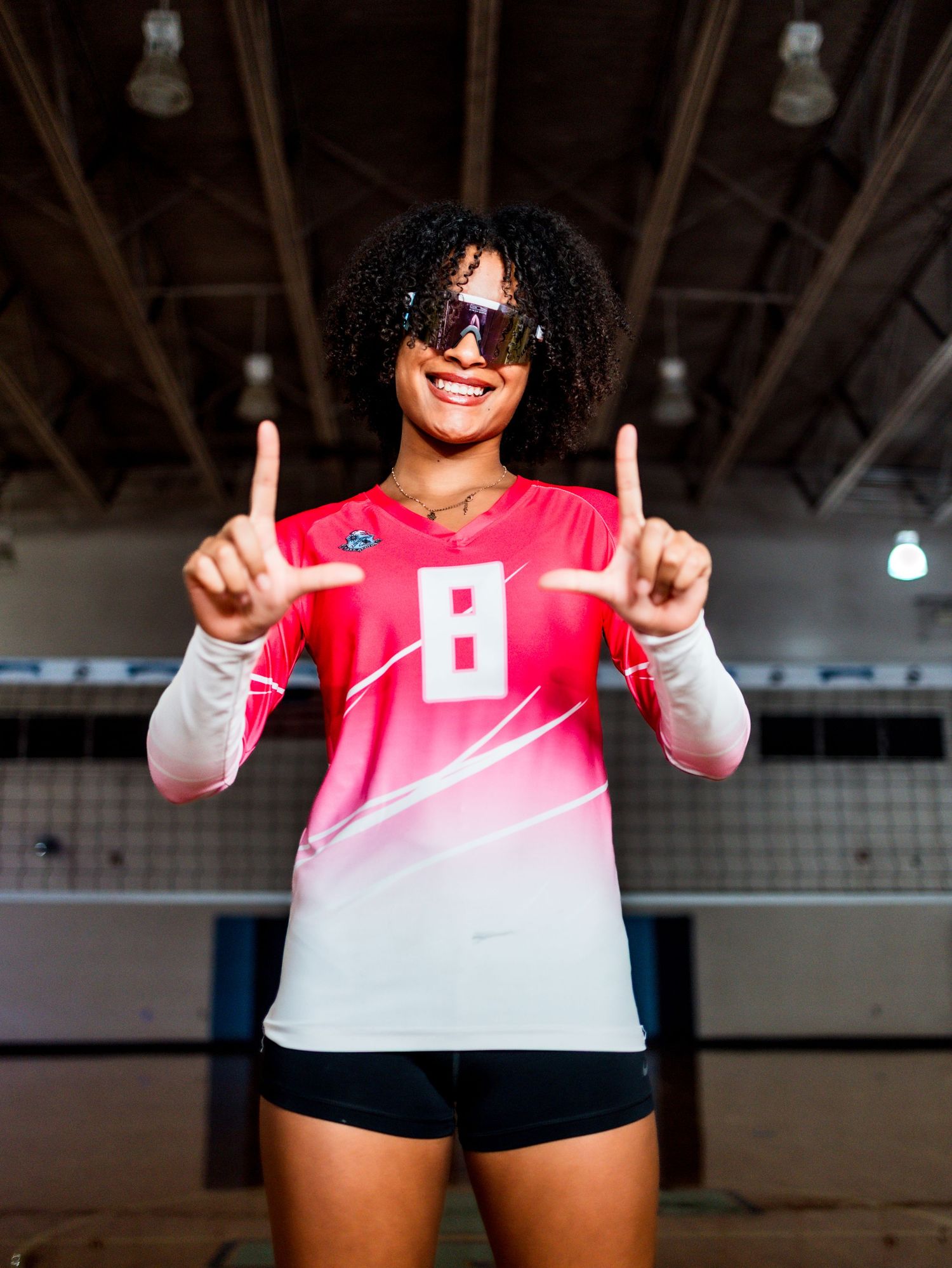 Volleyball player in pink jersey makes victory pose with upward pointing fingers in indoor gym.