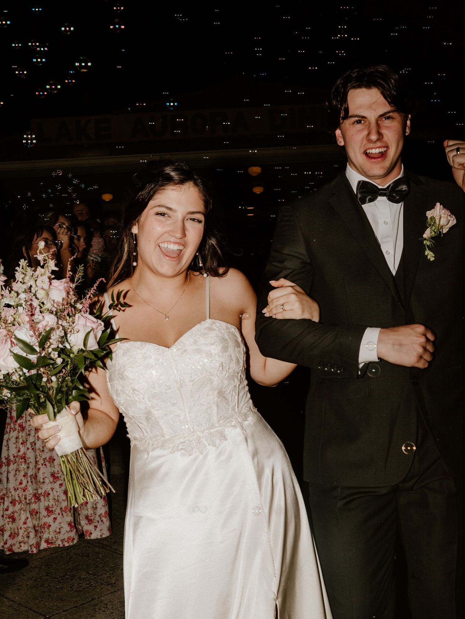 A couple in formal attire dances together at a dimly lit wedding reception venue with other guests in the background.
