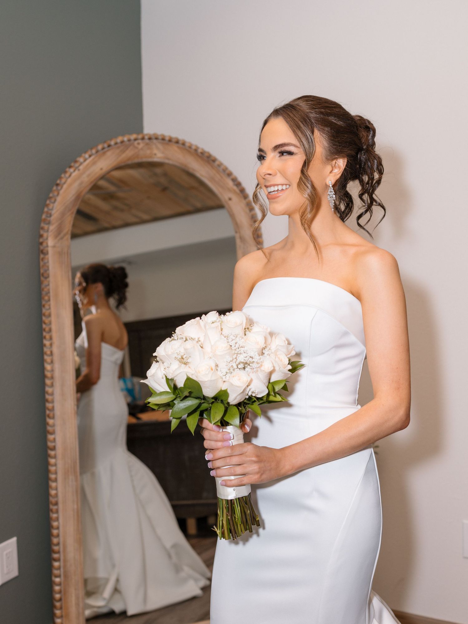 A bride in a white wedding dress holds a classic white bouquet while checking her reflection in an ornate mirror.