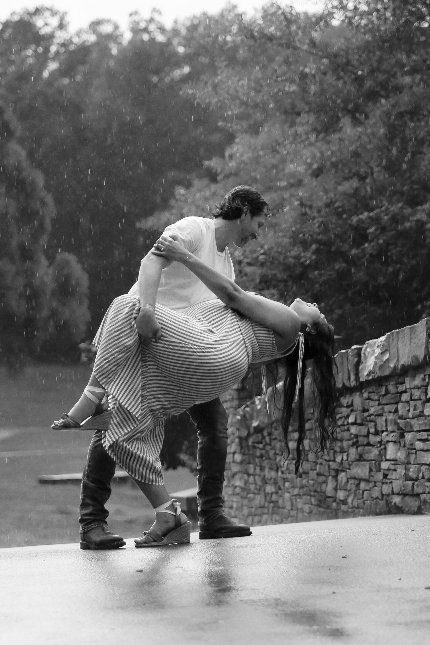 Couple dancing romantically in the rain, man dipping woman near stone wall, black and white photo.