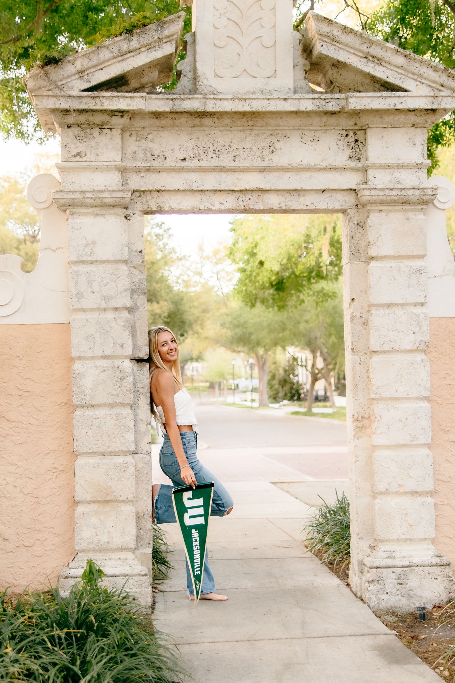 Young woman holding a JU pennant poses under a stone archway on a sunny day.