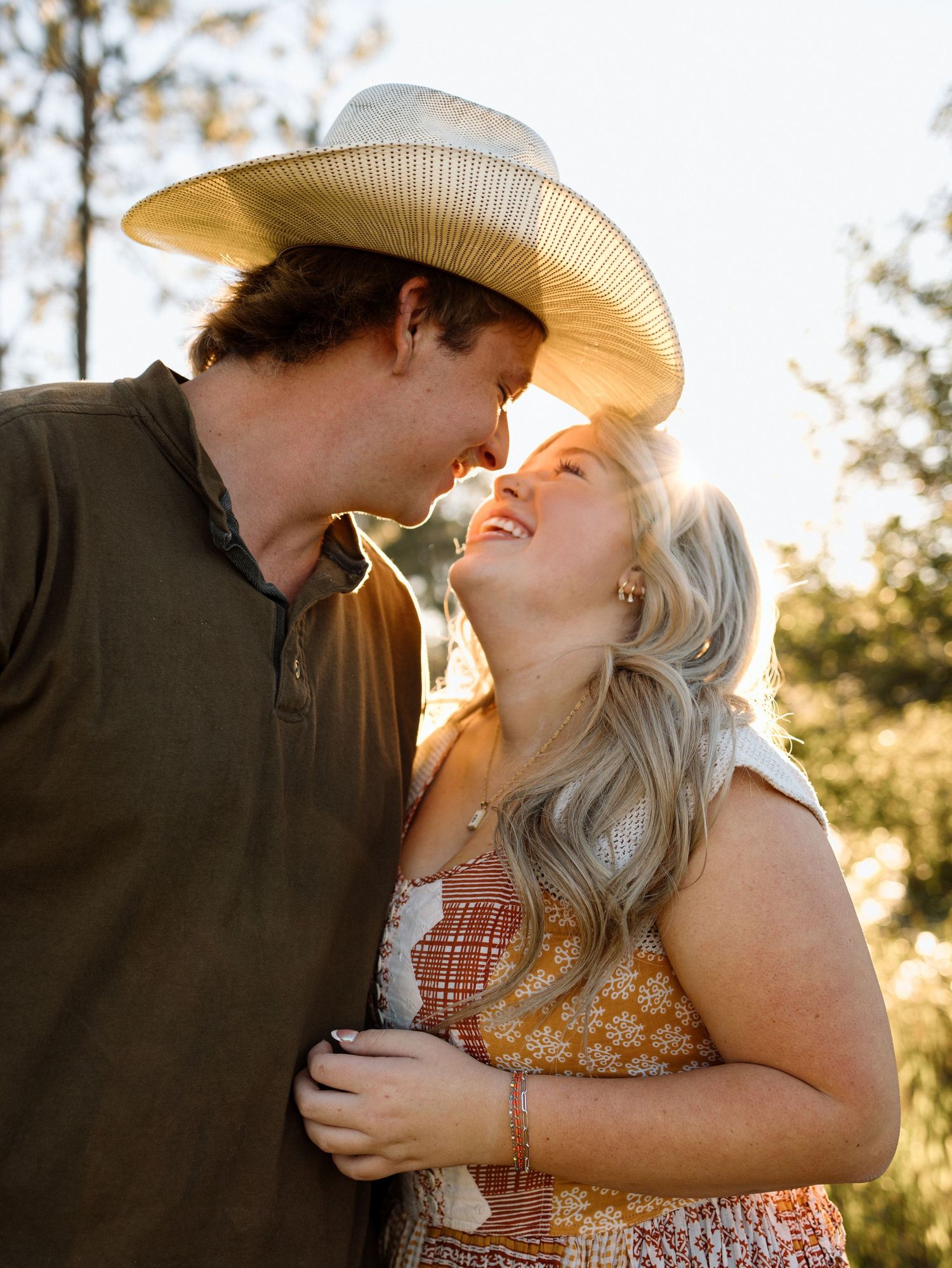 A couple in cowboy hats share a tender kiss in golden sunlight during a romantic outdoor photoshoot.