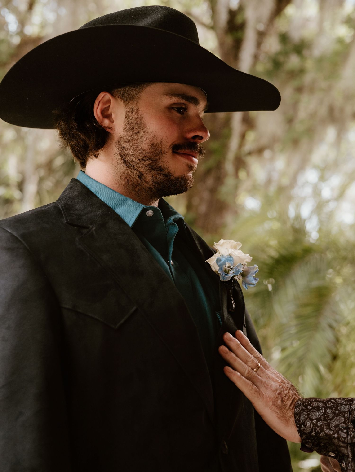 Groom in western attire with cowboy hat and boutonniere in natural setting.