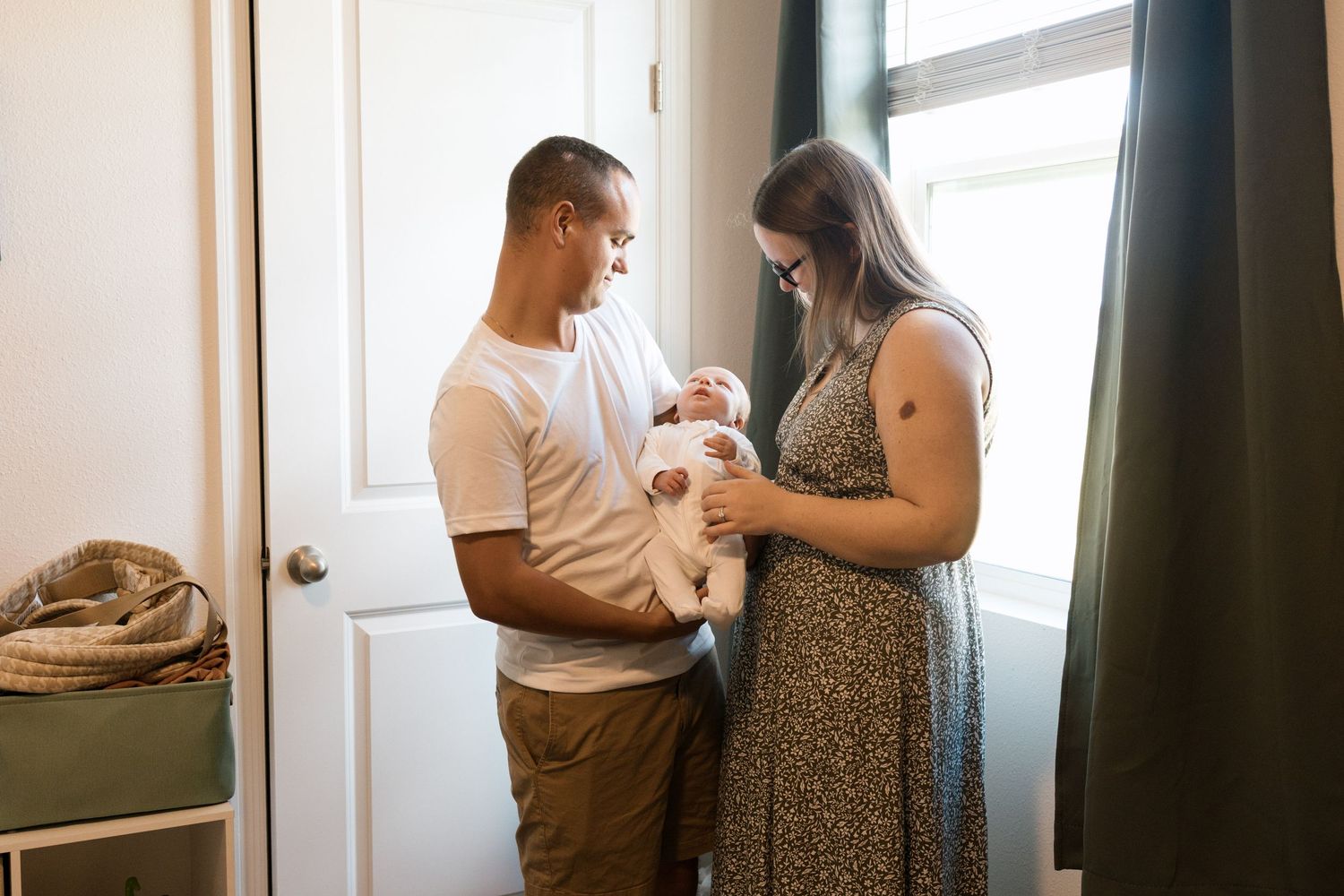 New parents stand together holding their newborn baby near a window in a warmly lit home.