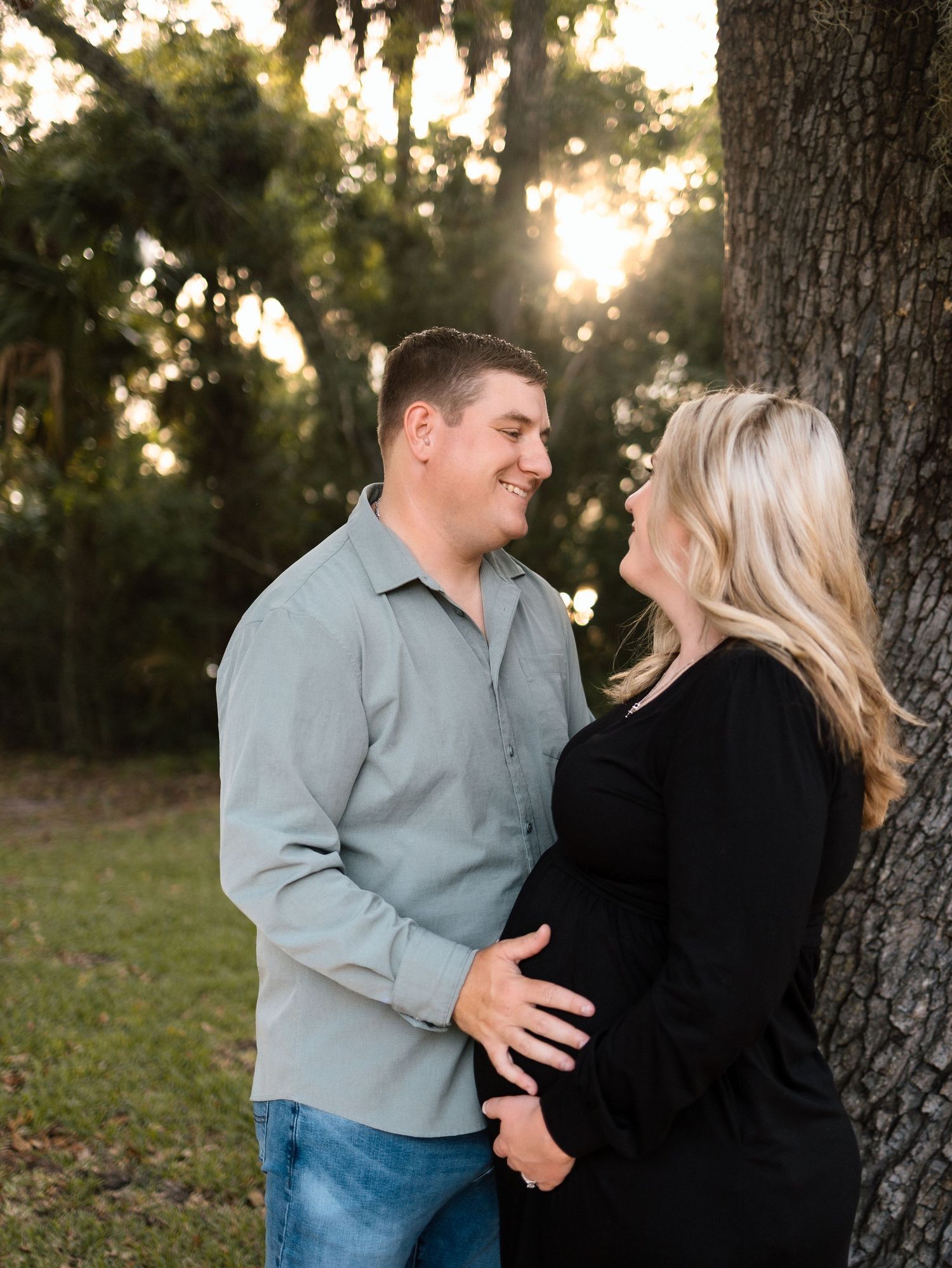 Couple sharing a tender moment by a tree at golden hour, man smiling at pregnant woman in black dress outdoors.