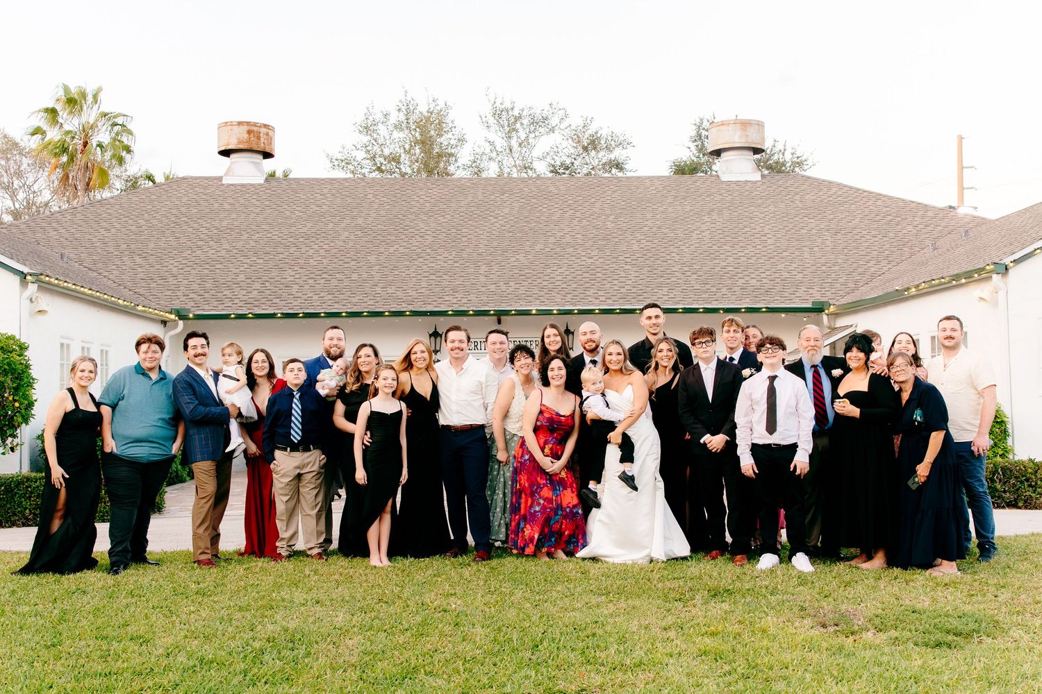 Large wedding party group photo outdoors in front of white building on green lawn.