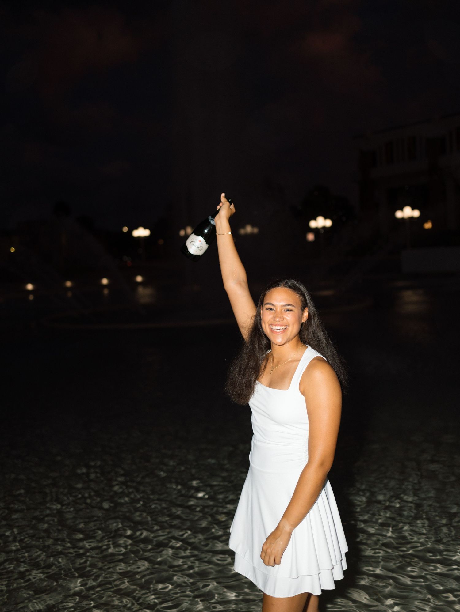 Young woman in white dress holding a white rose overhead, smiling at night outdoors.