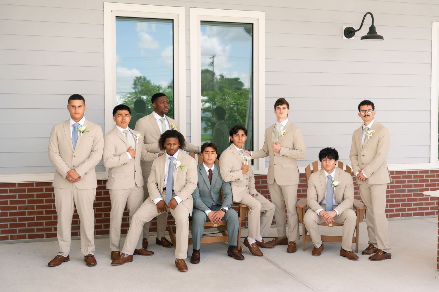 Nine groomsmen in tan suits pose together outside a white barn venue for a wedding party portrait.