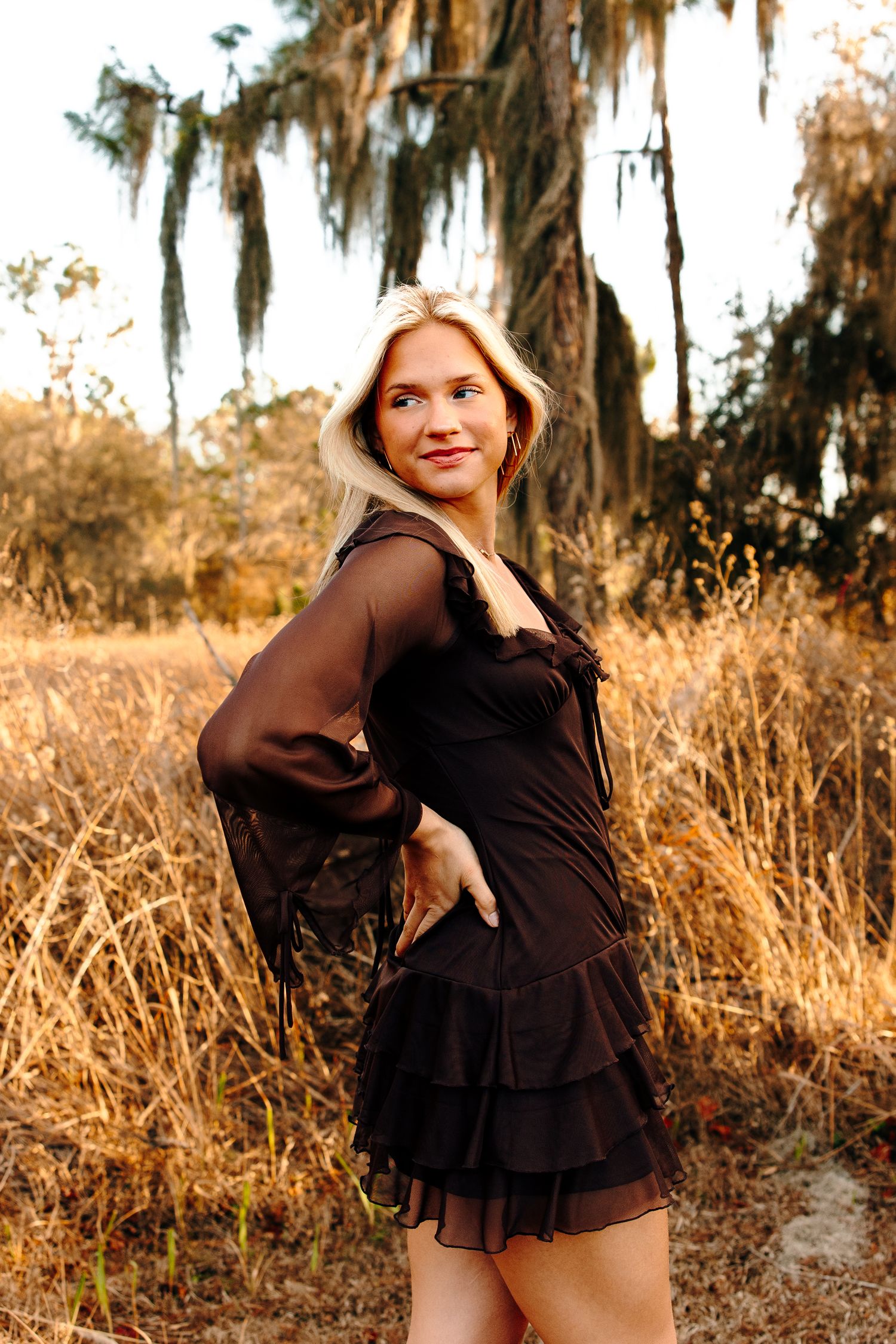 Blonde woman in black ruffled mini dress posing in golden field with Spanish moss trees backdrop.