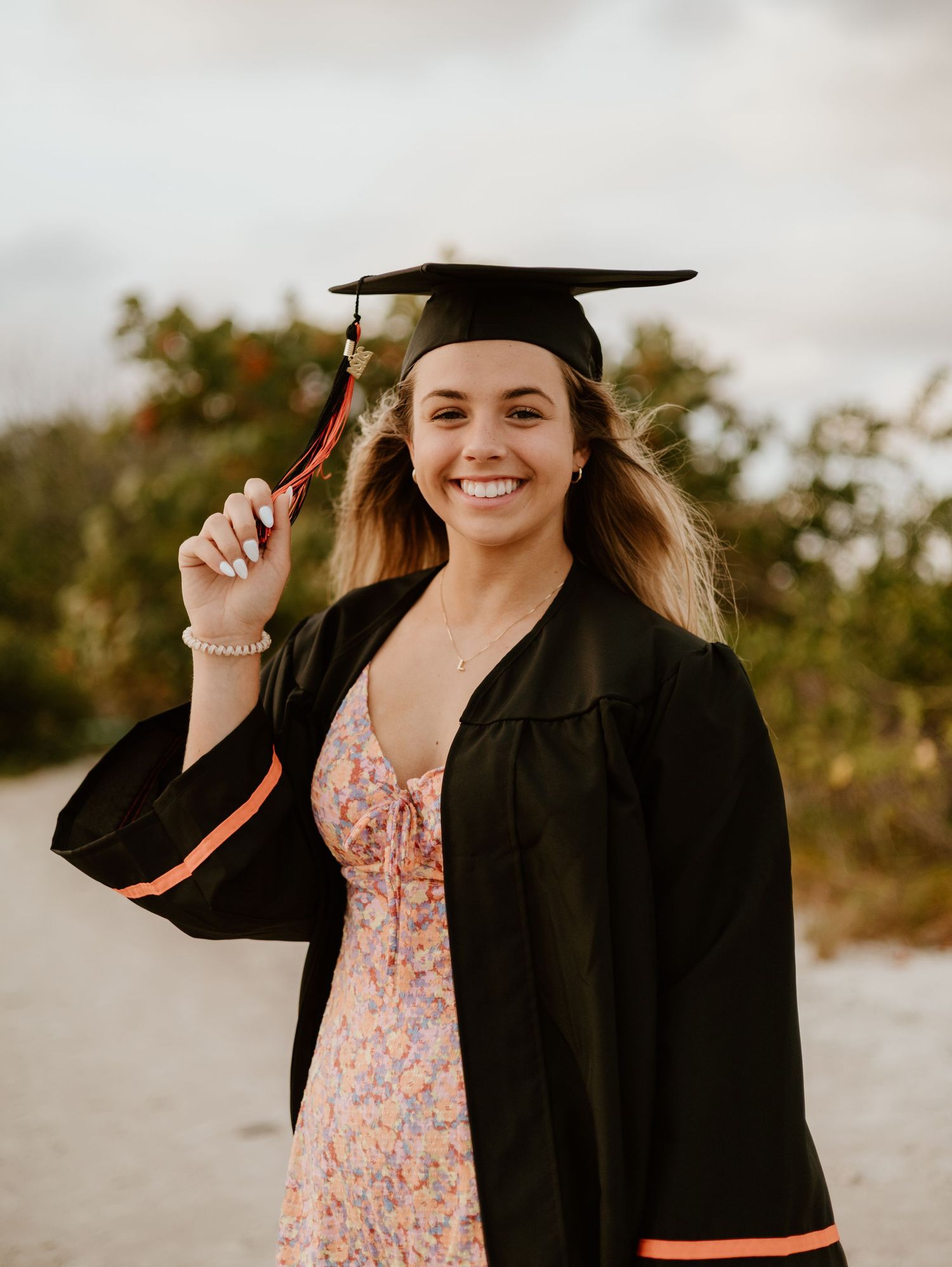 Graduate wearing black cap and gown with floral dress underneath poses outdoors at sunset for graduation photos.