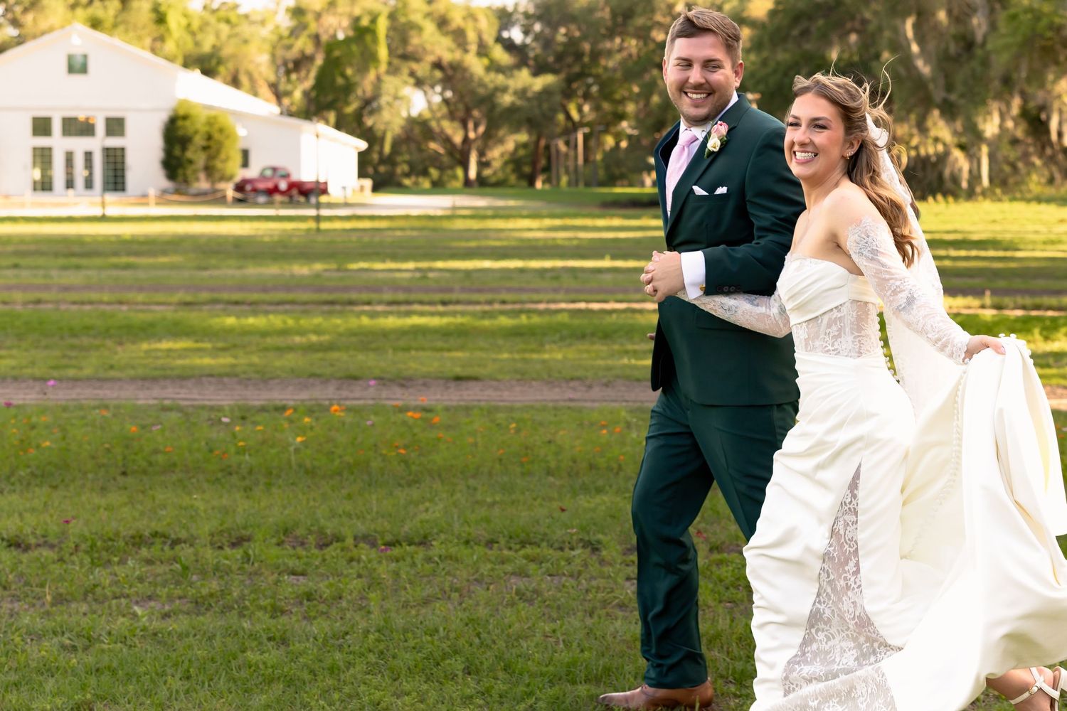 Joyful bride and groom in green tuxedo walking on lush lawn at outdoor wedding venue on sunny day.