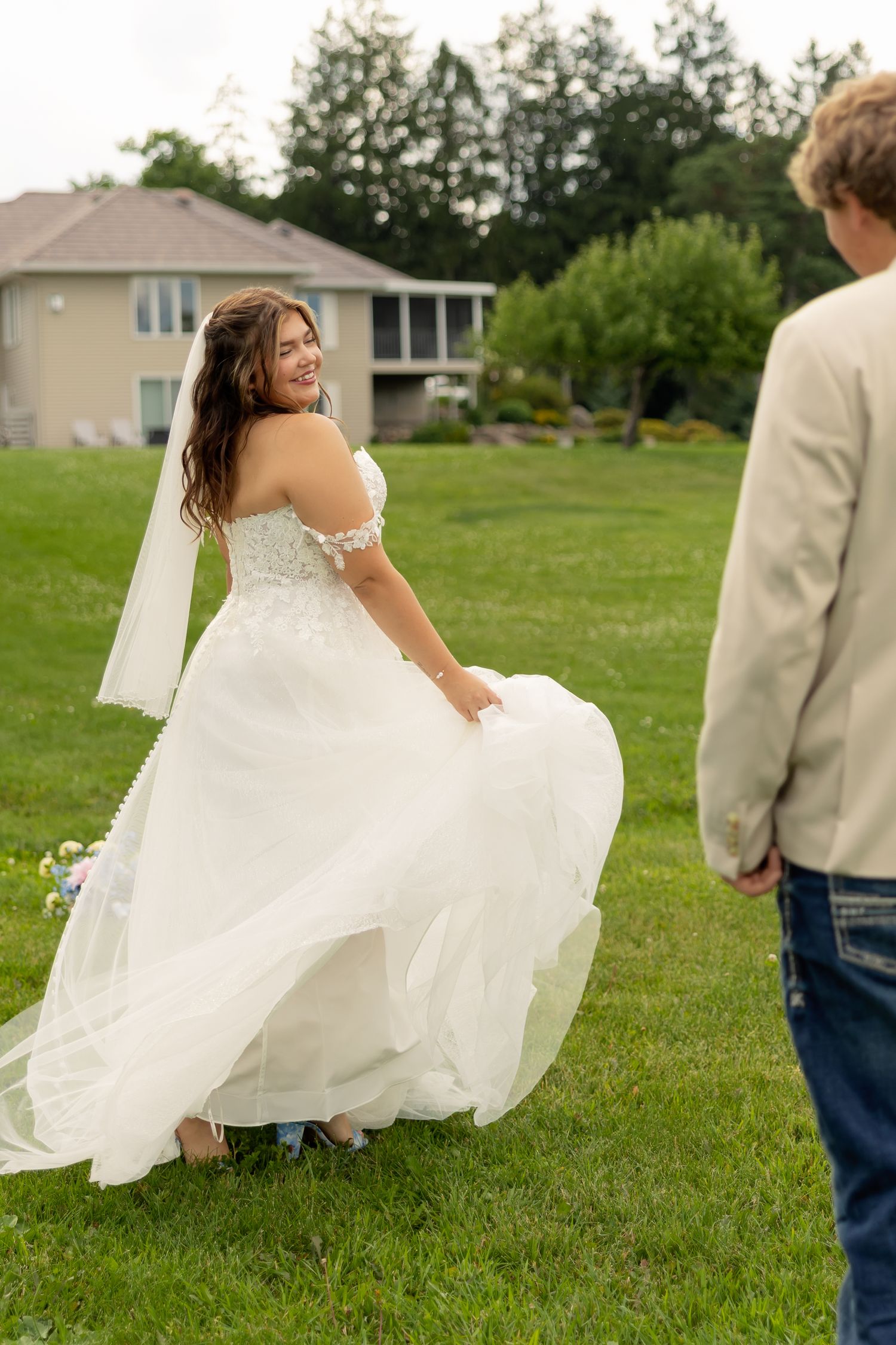 Smiling bride in white off-shoulder gown twirling on green lawn, looking back at groom in tan jacket.