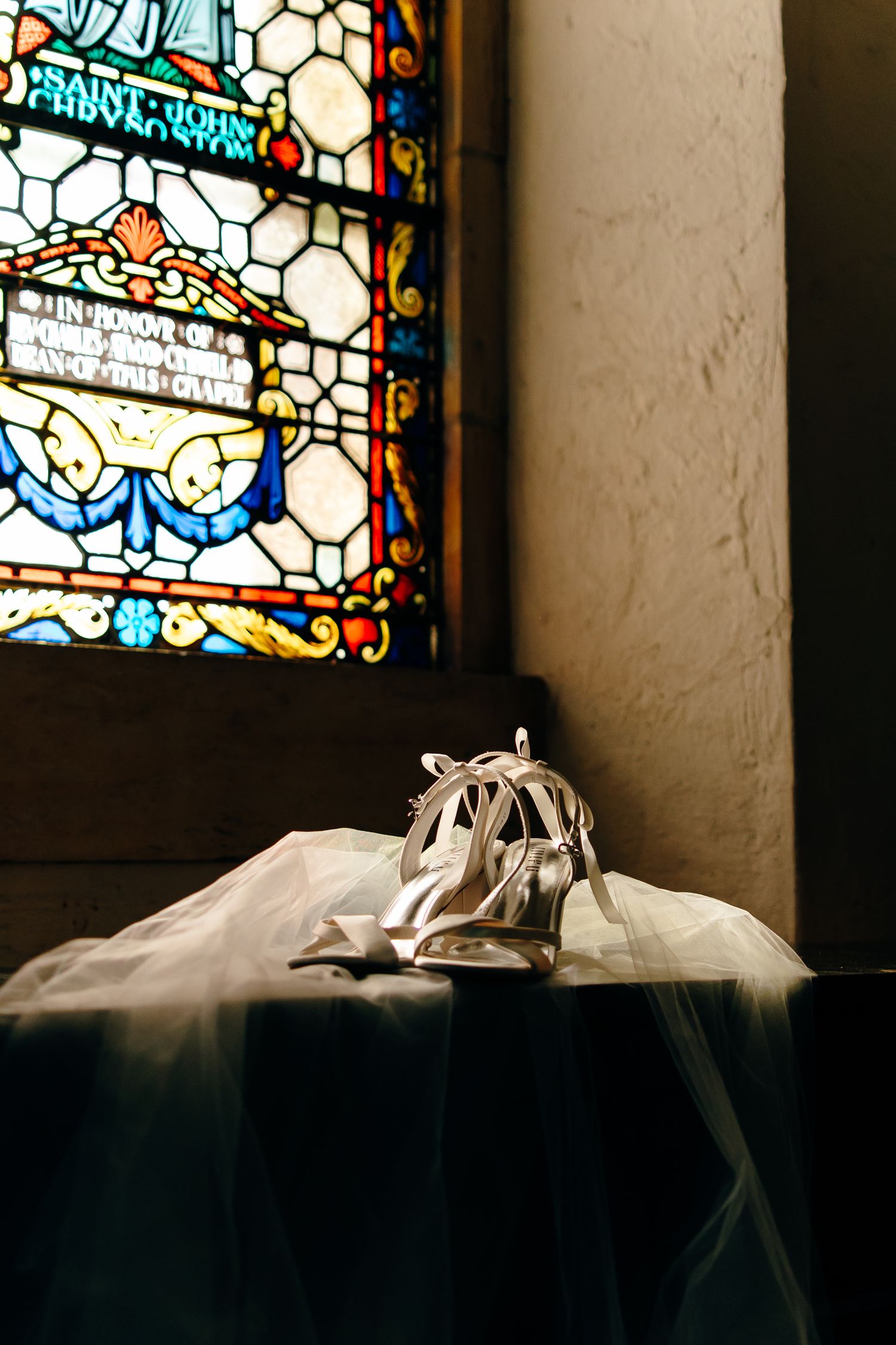 Bridal heels with ribbon ties rest on white tulle fabric before a colorful stained glass church window.