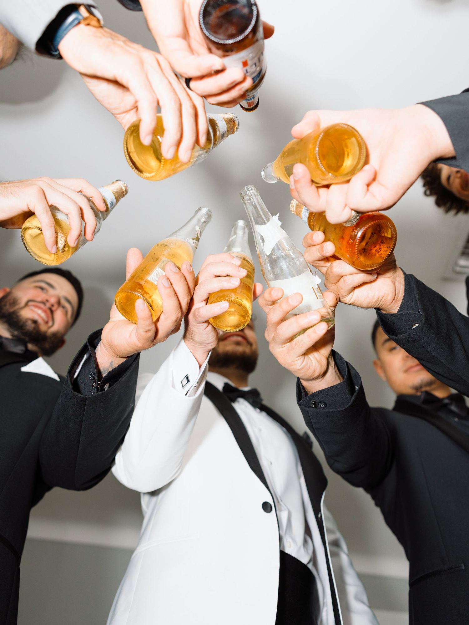 Groomsmen in tuxedos toasting with beer bottles at a wedding celebration.
