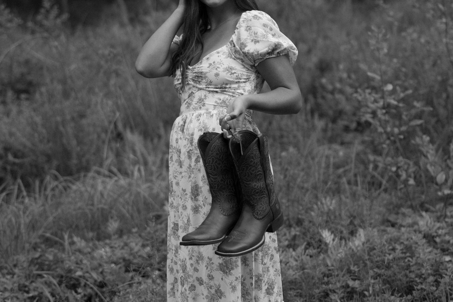 A high school girl in a floral dress walks through a lush summery garden with rustic wooden fencing in the background holding cowboy boots.