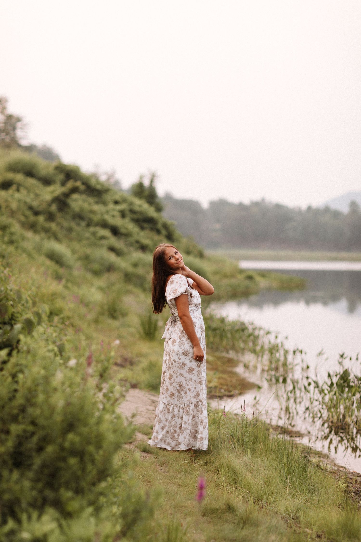 A person in a floral dress poses candidly by a serene lakeside meadow in summer Surry Lake, NH