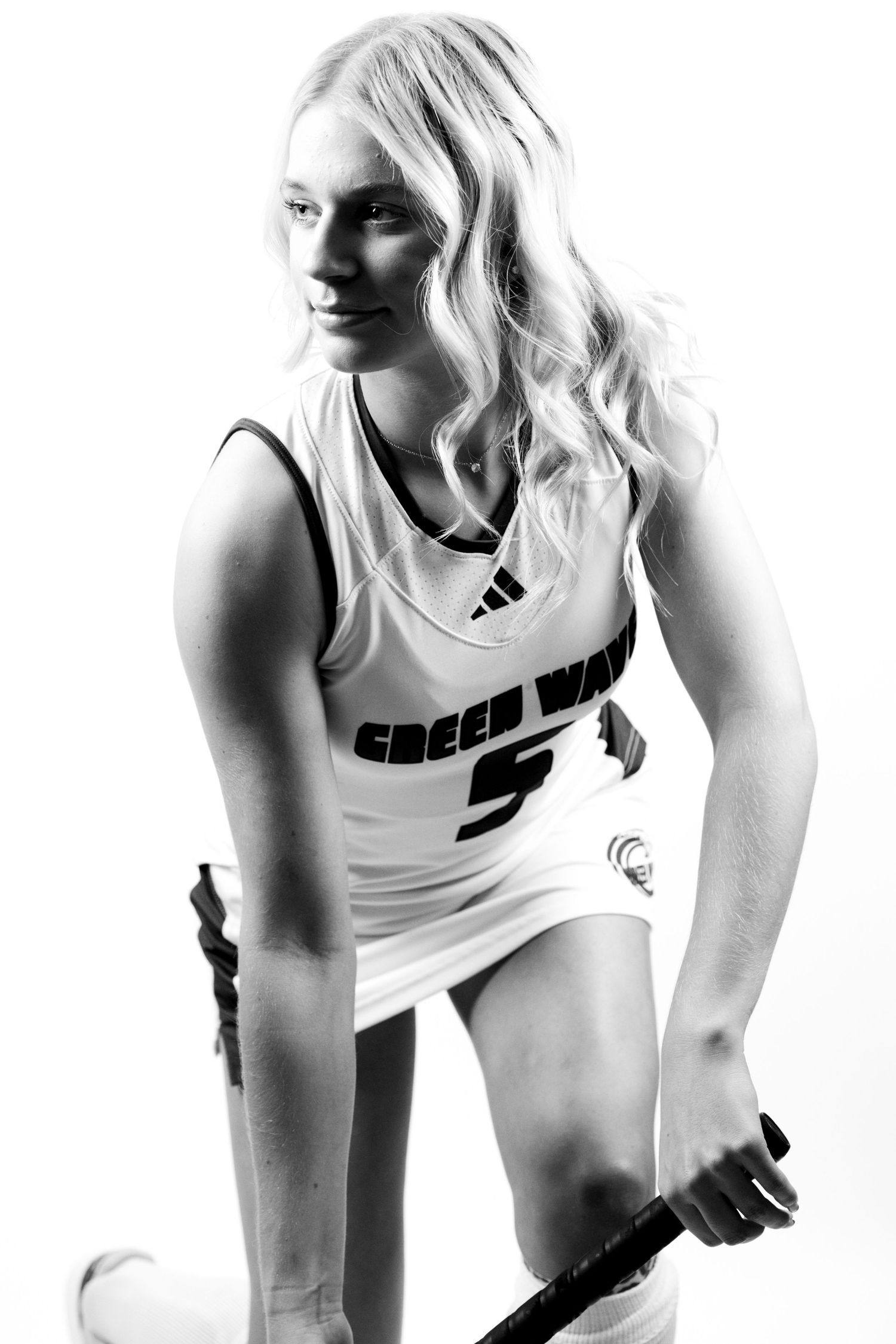 Black and white portrait of a field hockey player in a white jersey kneeling while holding a hockey stick during senior photoshoot.
