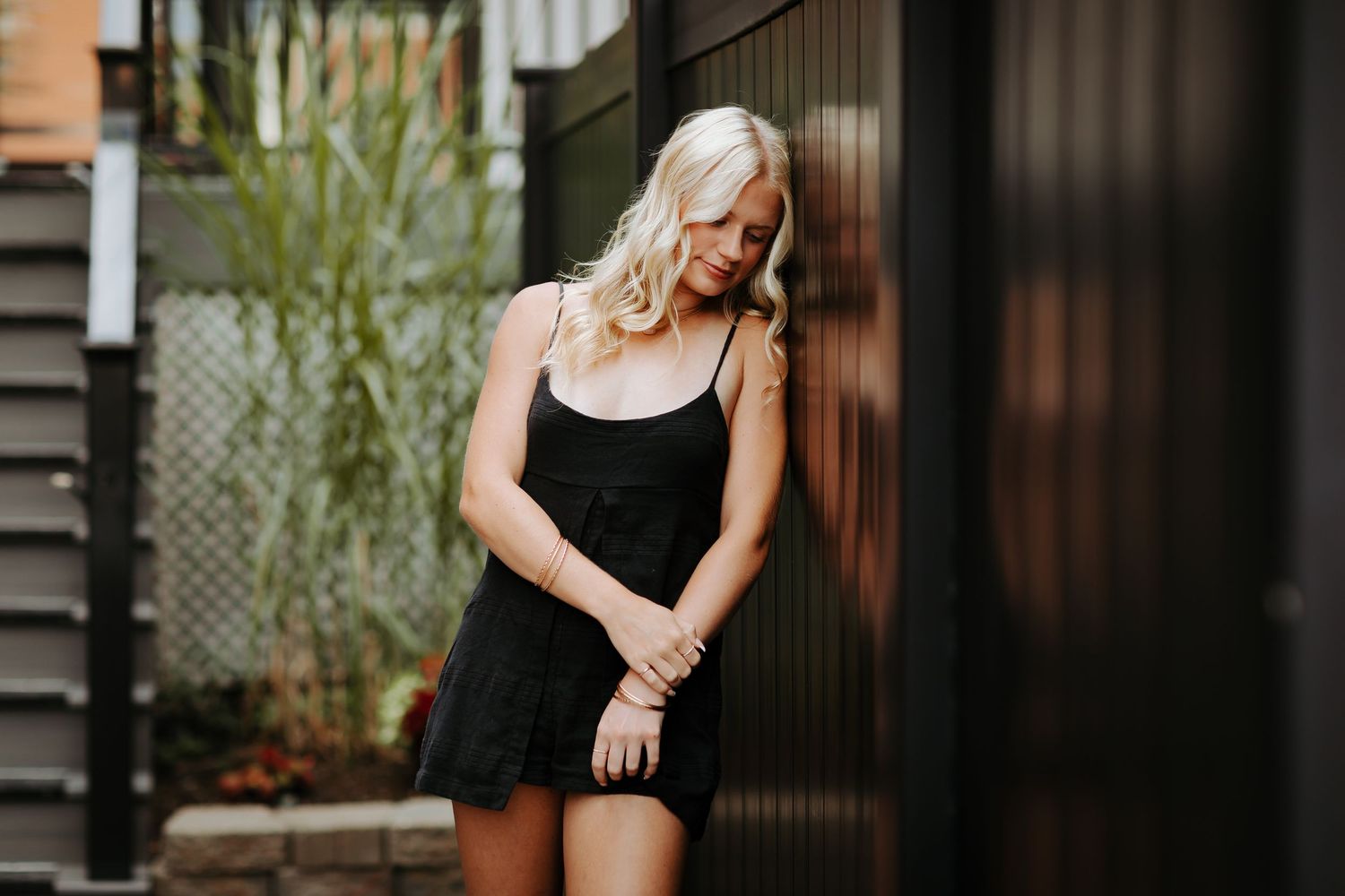 Soft natural lighting falls on a high school girl in a black slip dress against a dark wooden wall with bamboo plants in the background.
