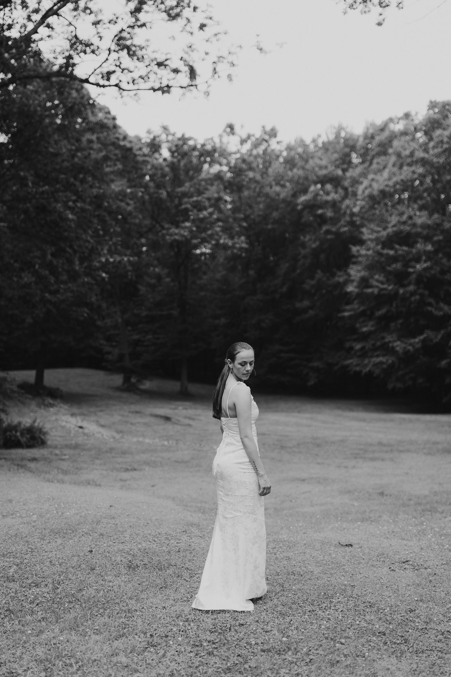 A high school girl in a flowing white dress stands in a grassy meadow surrounded by trees in black and white NH