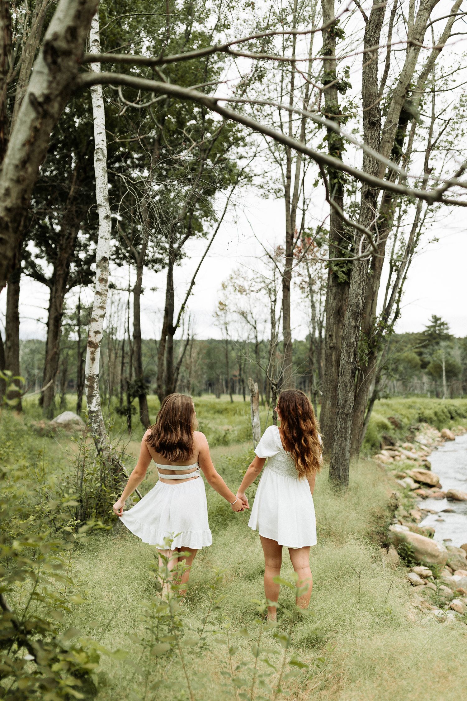 Best friends in white dresses run playfully through a wooded area with sunlit trees and grass in the background for a senior photoshoot NH