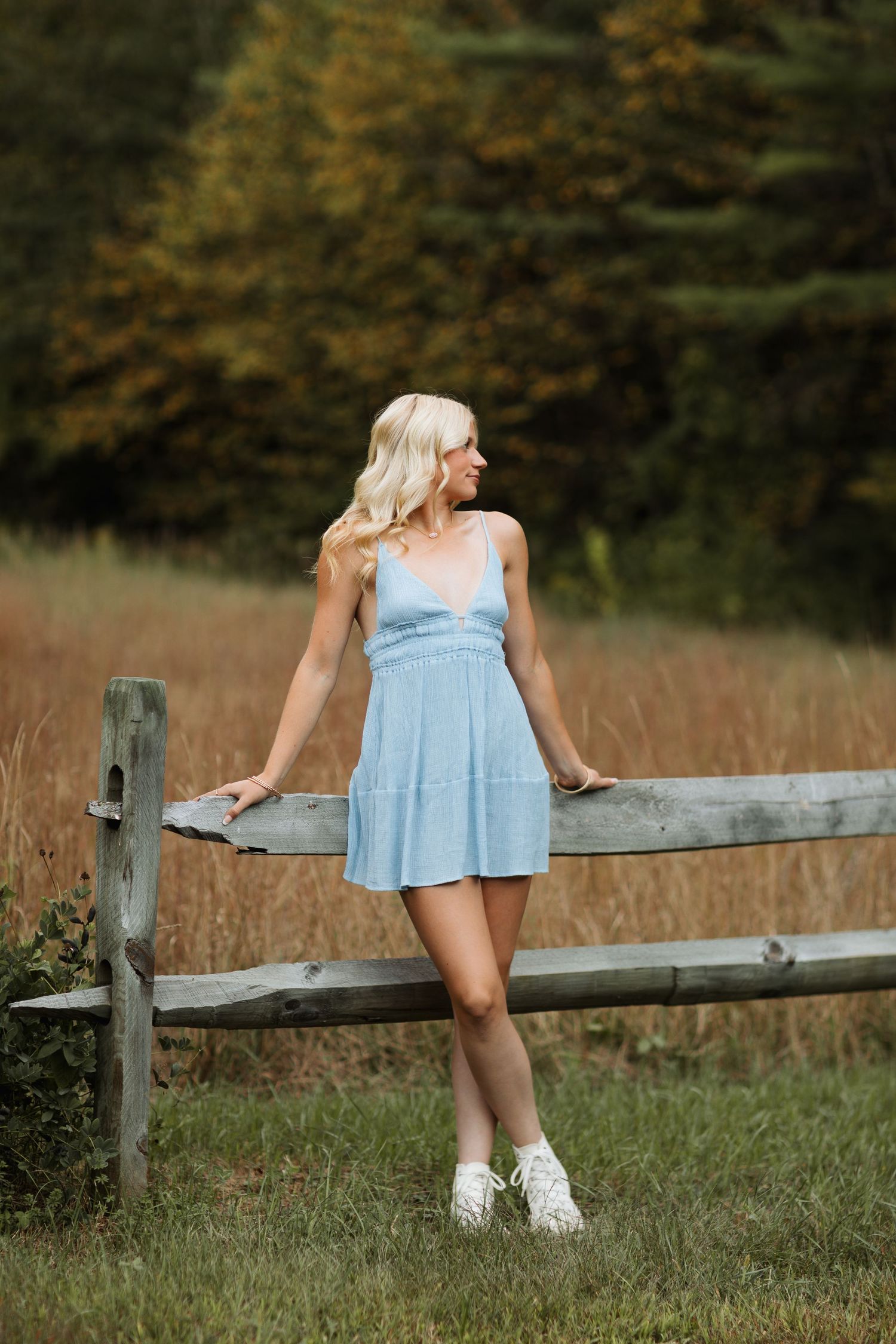 A high school girl in a light blue summer dress leans against a wooden fence post in a serene field during autumn in Swanzey, New Hampshire.