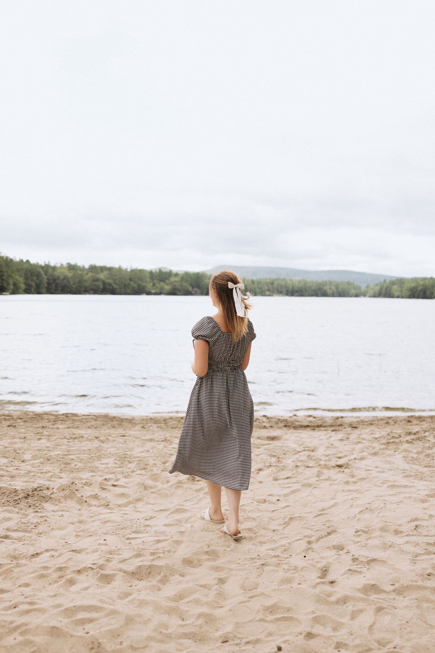 Someone in a striped gray dress stands barefoot on a sandy beach looking out at a serene lake surrounded by trees, NH, Portsmouth