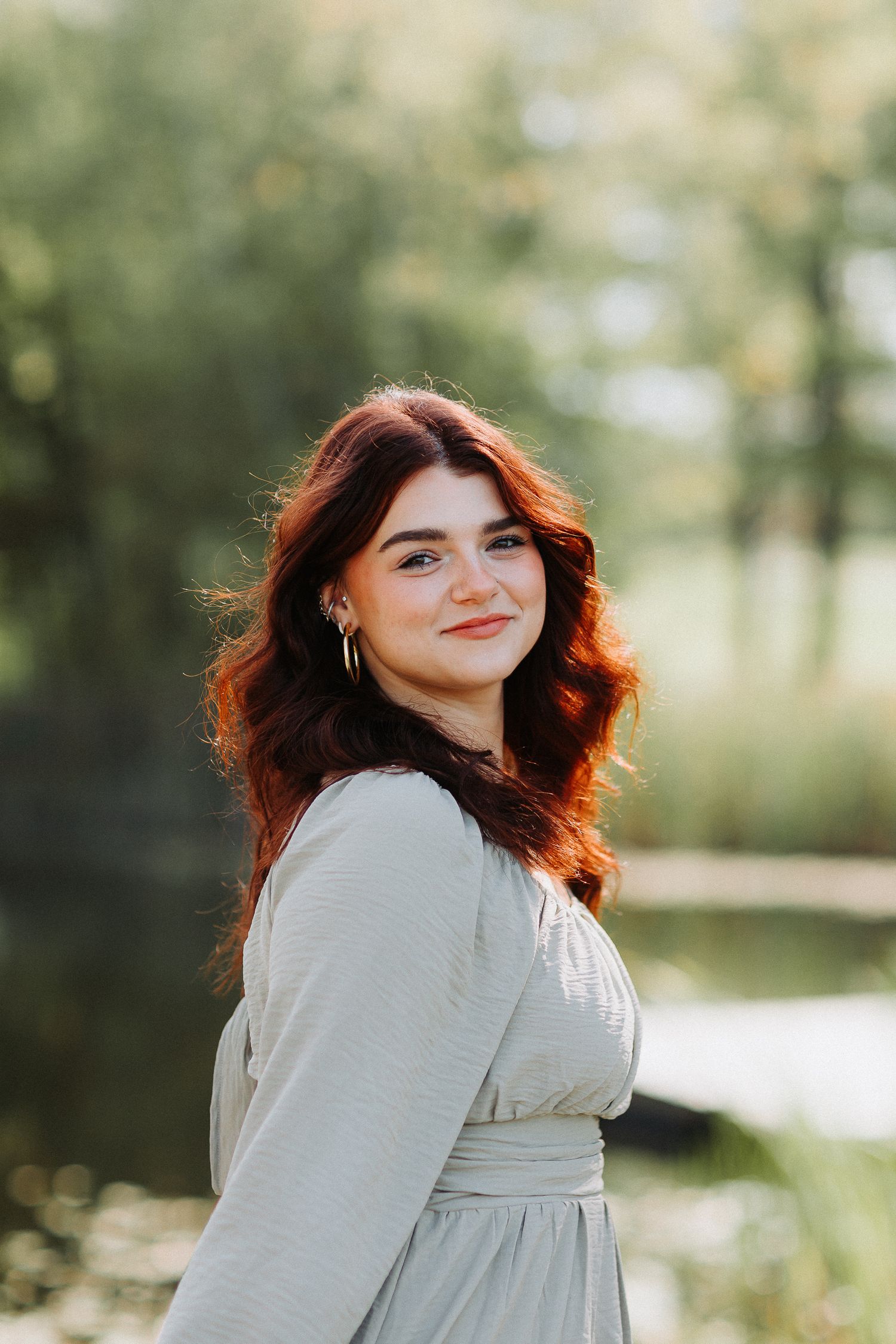 A person with wavy red hair wearing a light-colored dress poses outdoors with sunlit greenery in the background. Alysons Orchard NH