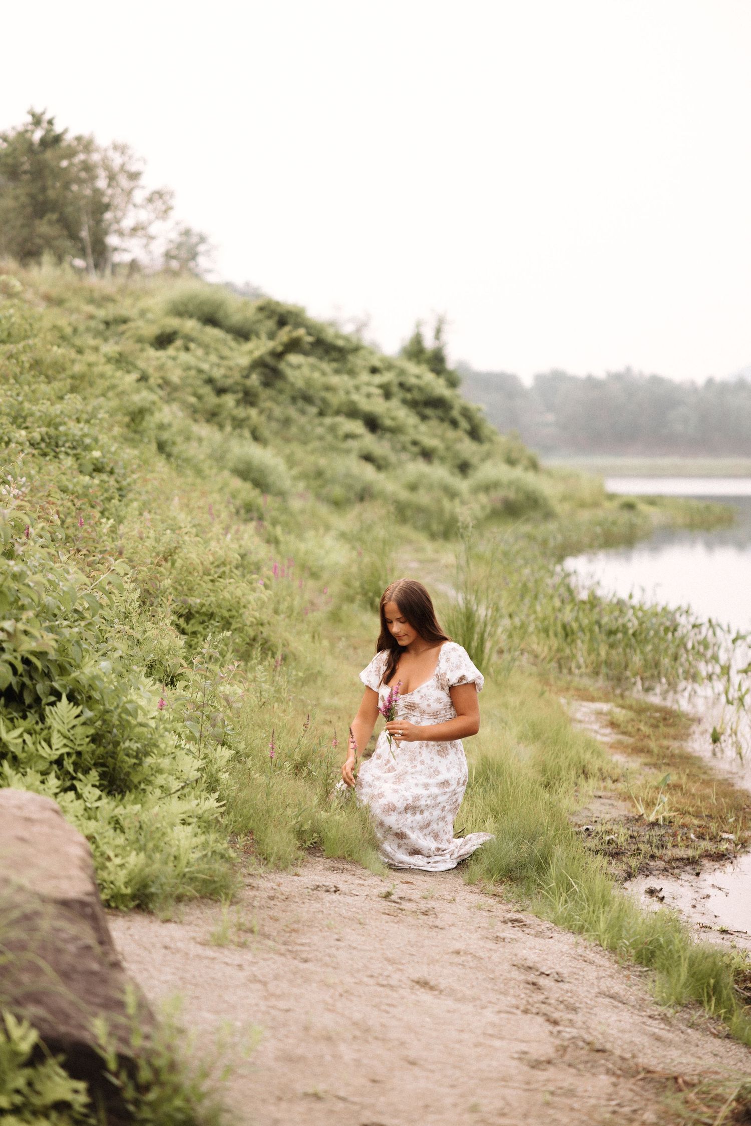 A girl in a white dress sitting by a riverside on a sunny summer day with lush greenery in the background Keene, New Hampshire