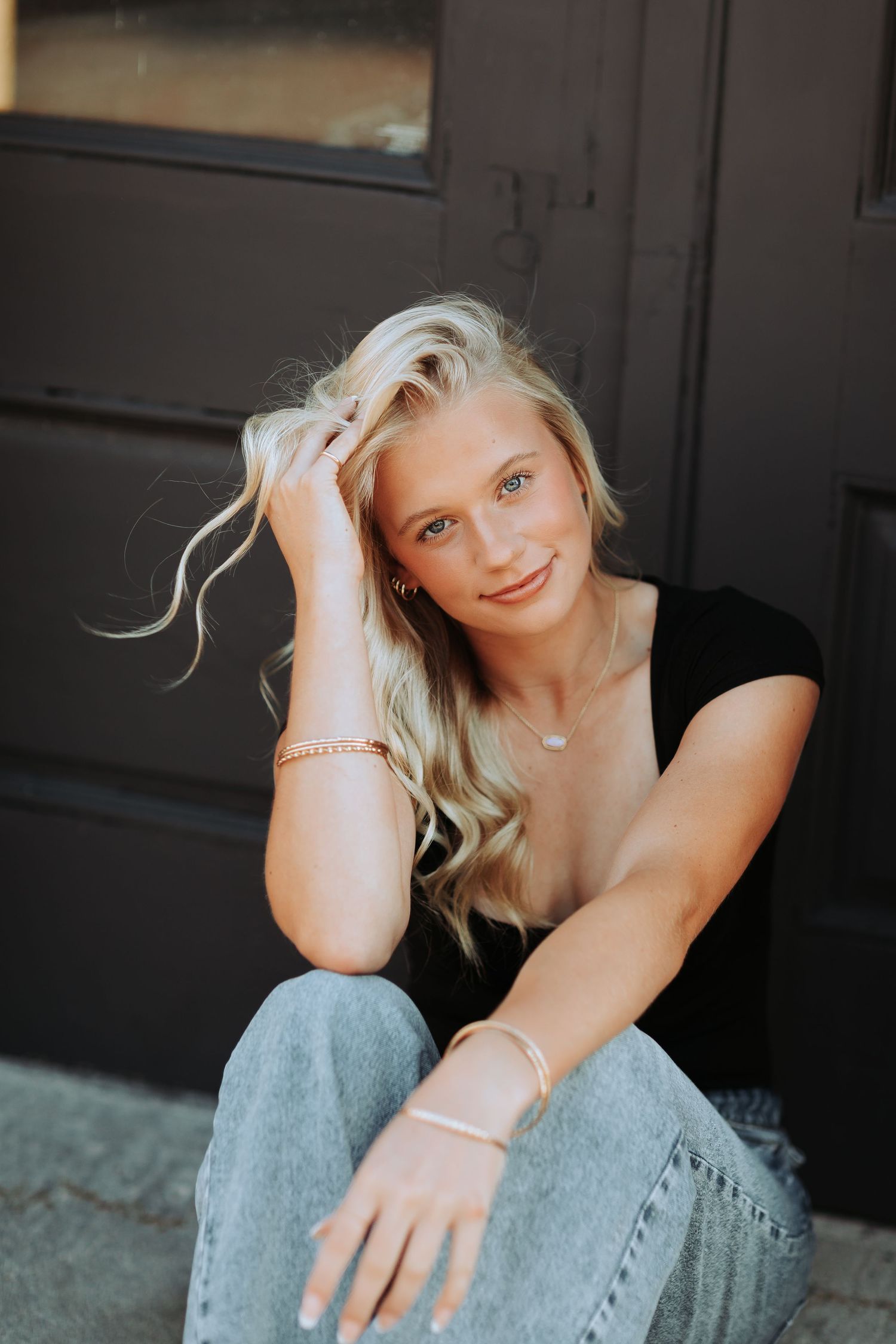 A lifestyle portrait of a young woman with blonde hair wearing a black top and jeans, sitting casually outdoors. New Hampshire Senior Photo