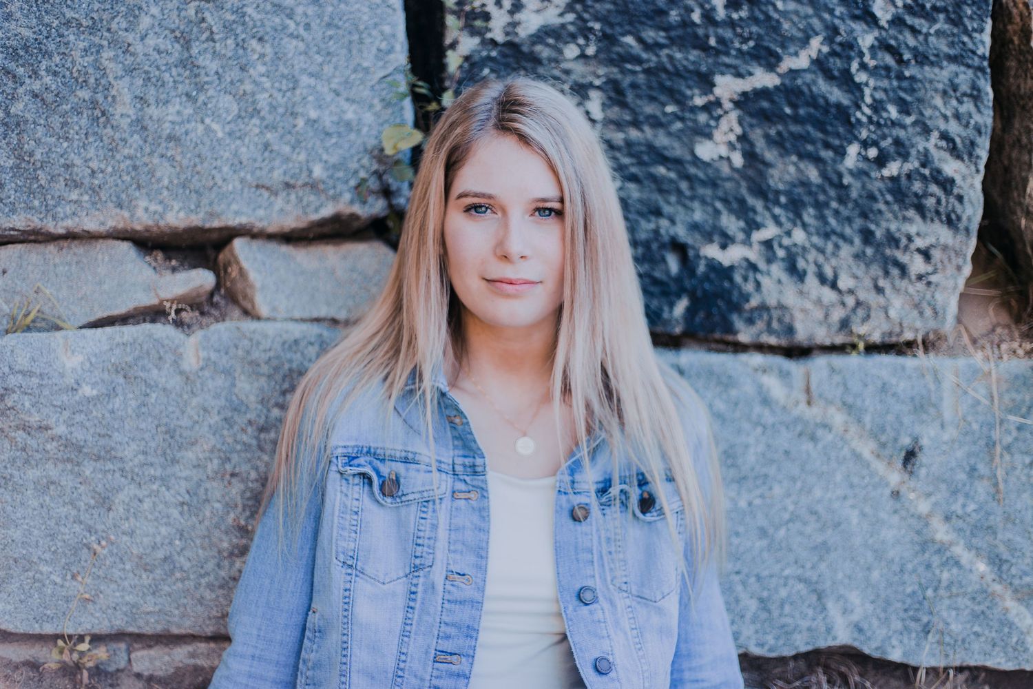 A high school senior with long blonde hair wearing a denim jacket poses against a stone wall background at Ashuelot Park in Keene, NH