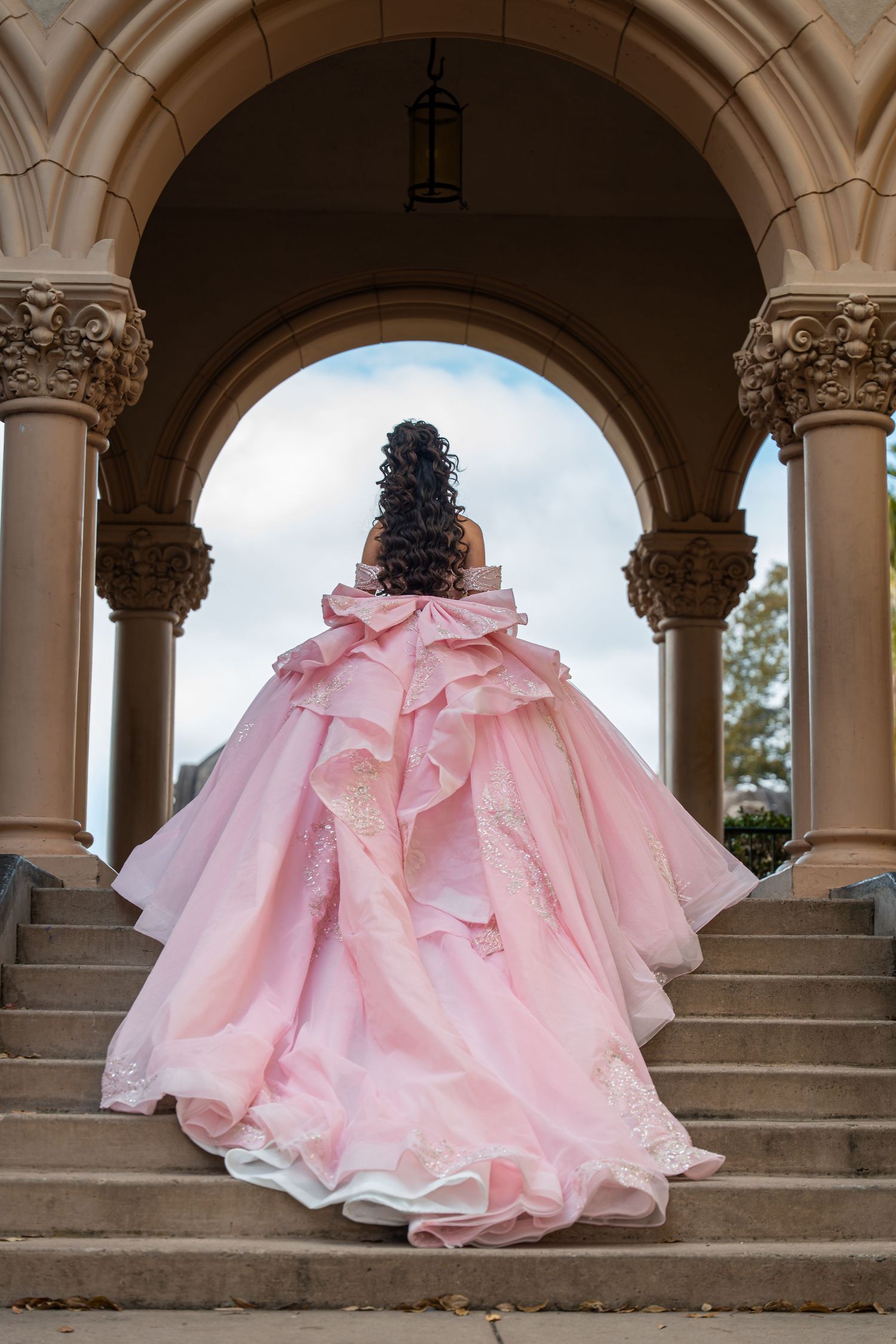 Young woman in elegant pink ball gown with large bow stands on stone steps under ornate arch.