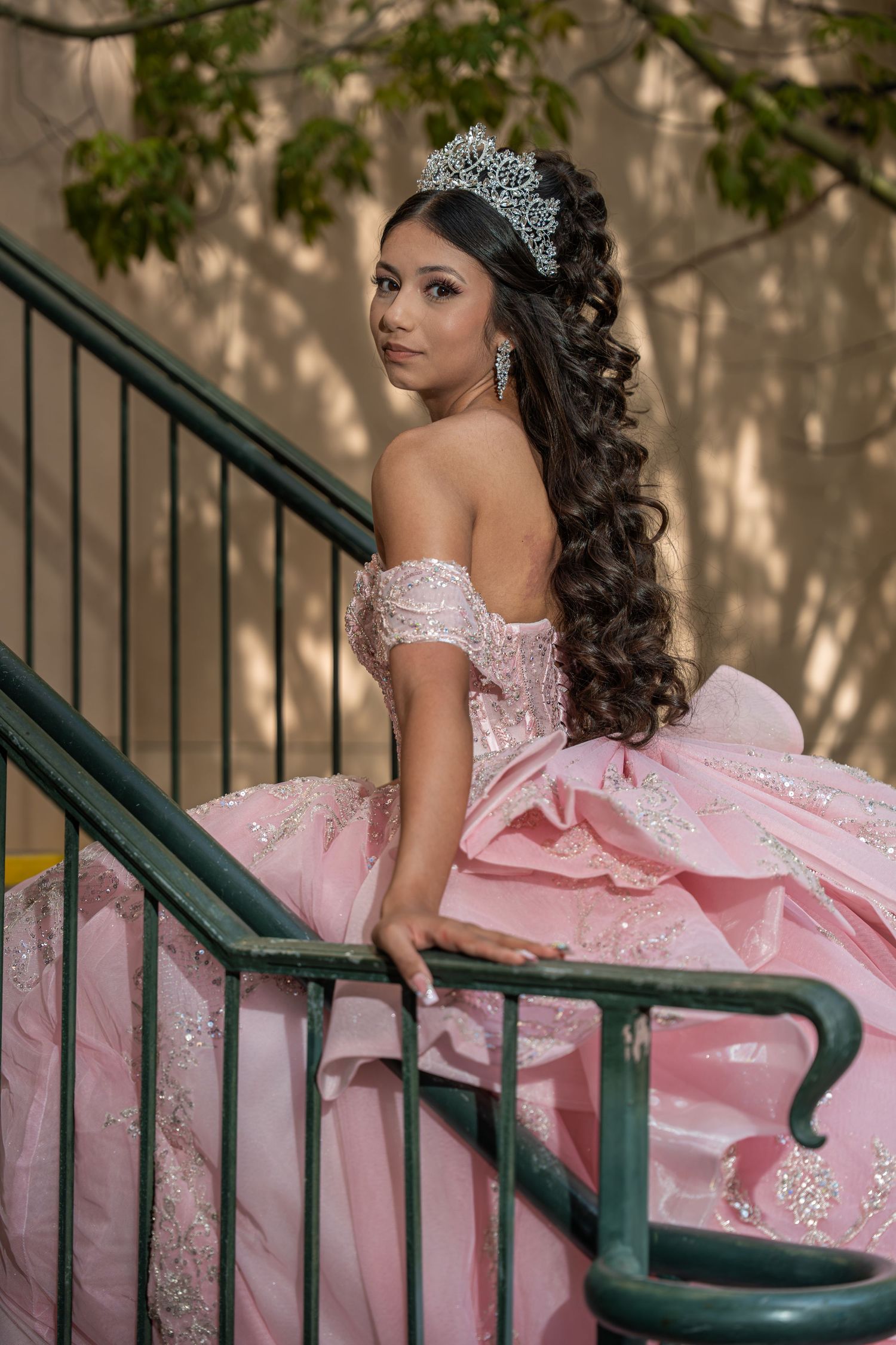 Young Latina in pink quinceañera ball gown with silver tiara poses gracefully on outdoor staircase.