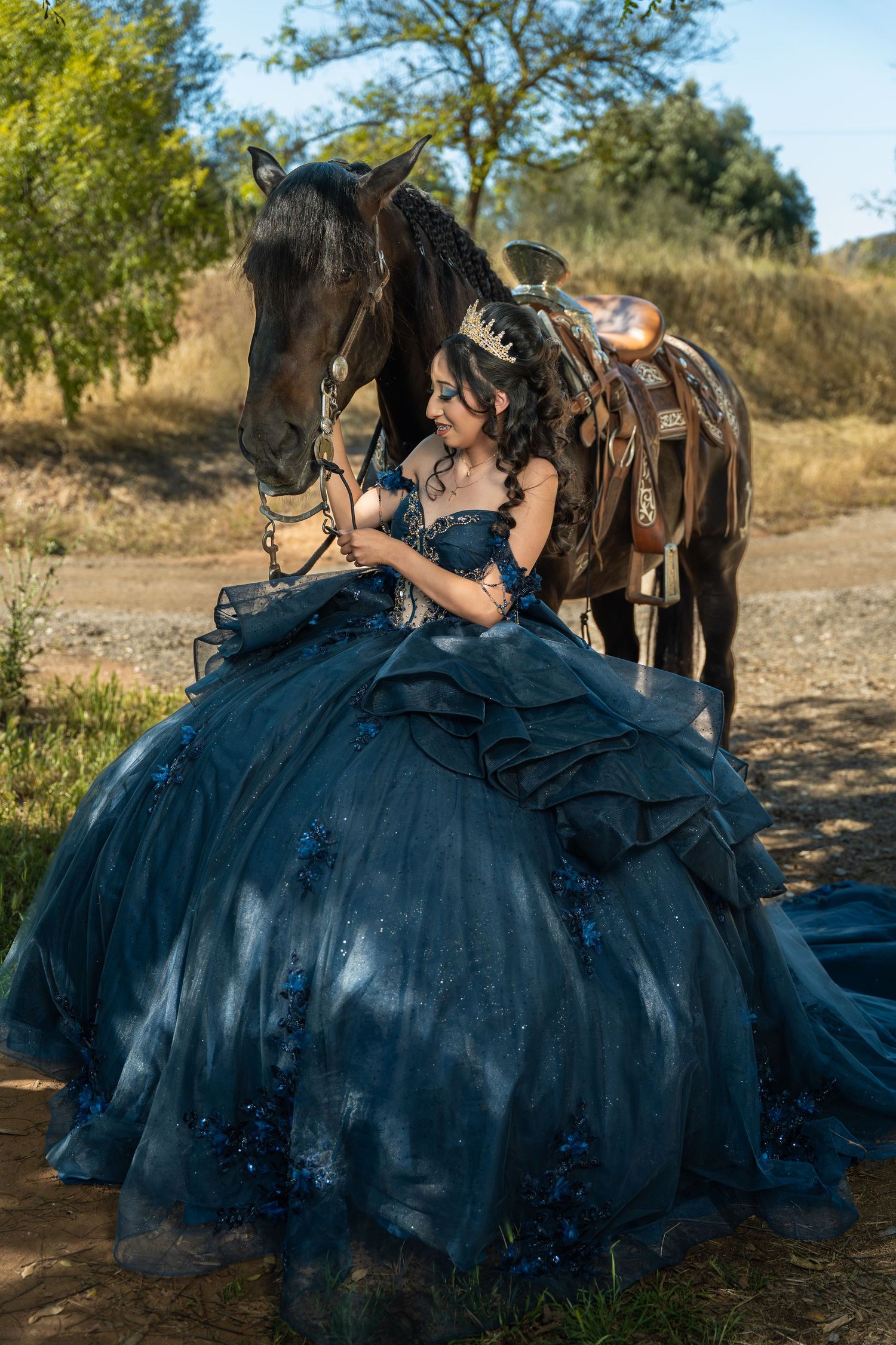Young woman in blue quinceañera ball gown smiling beside a dark horse in a sunny outdoor setting.