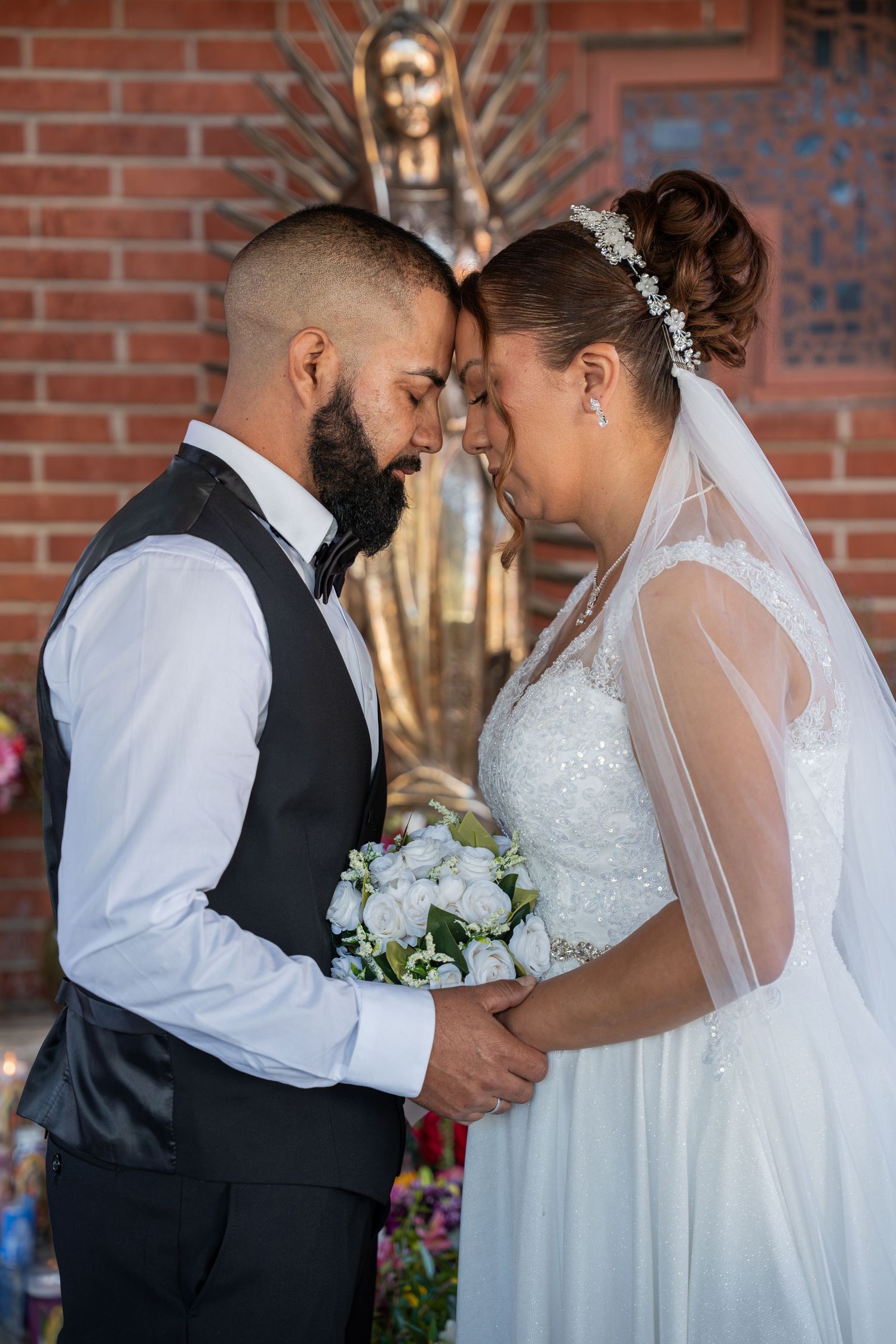 Bride and groom touching foreheads tenderly while holding a white bouquet in front of a brick church.