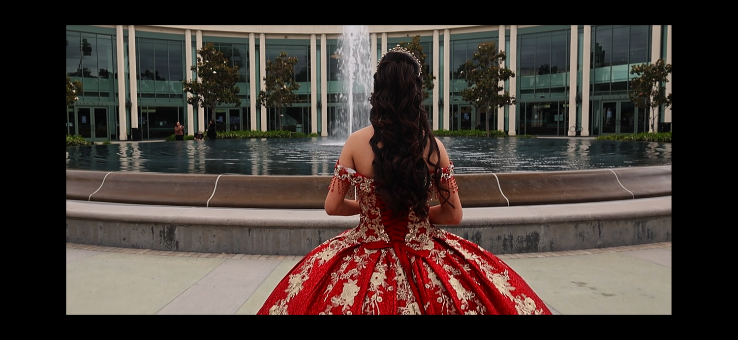 Young woman in red floral ball gown poses before elegant fountain in formal outdoor setting.