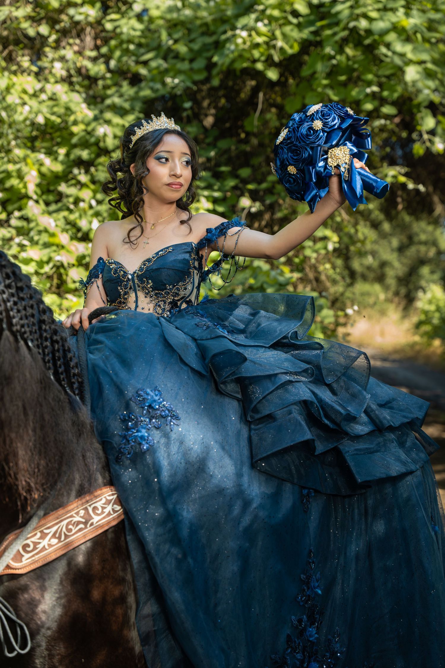 Young woman in navy blue quinceañera gown and crown sits on horseback holding blue floral bouquet outdoors.