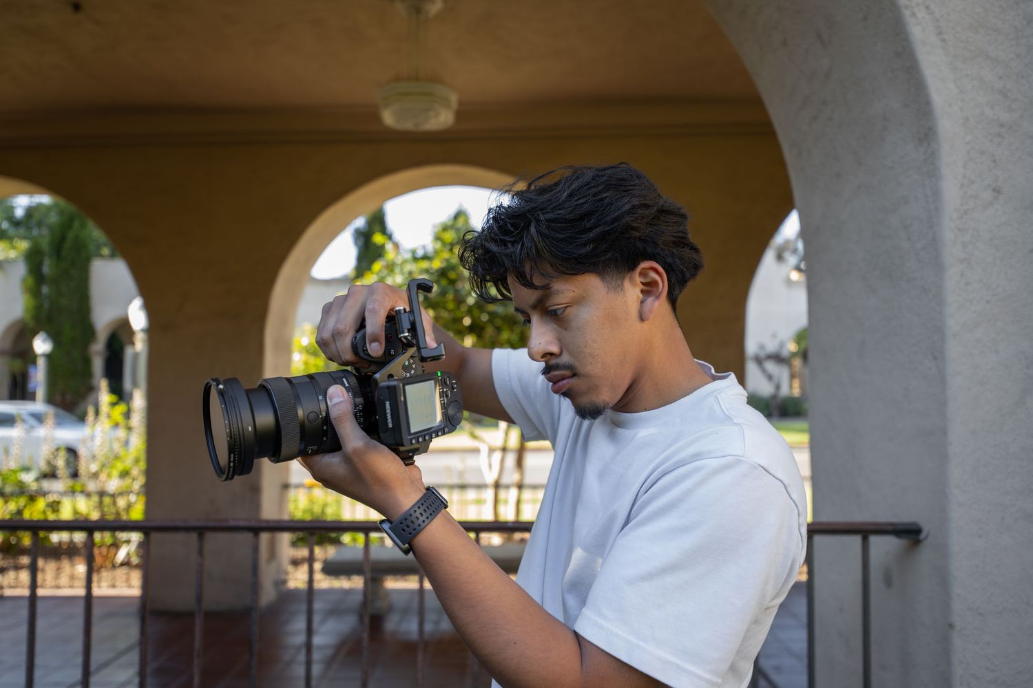 Young man with dreadlocks holding a DSLR camera outdoors, photographing near arched columns.