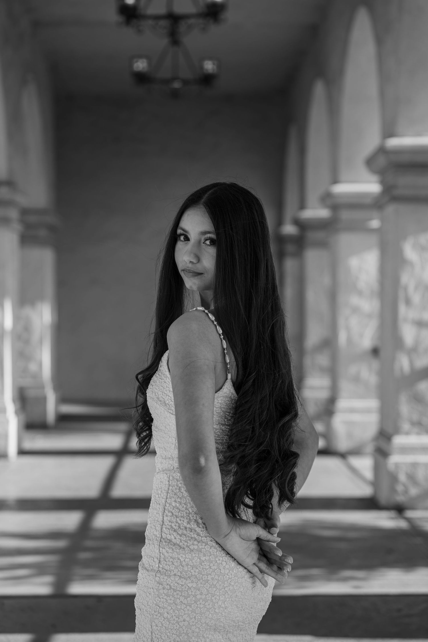 Young woman with long dark hair poses gracefully in a white dress inside an elegant columned hallway, black and white photo.