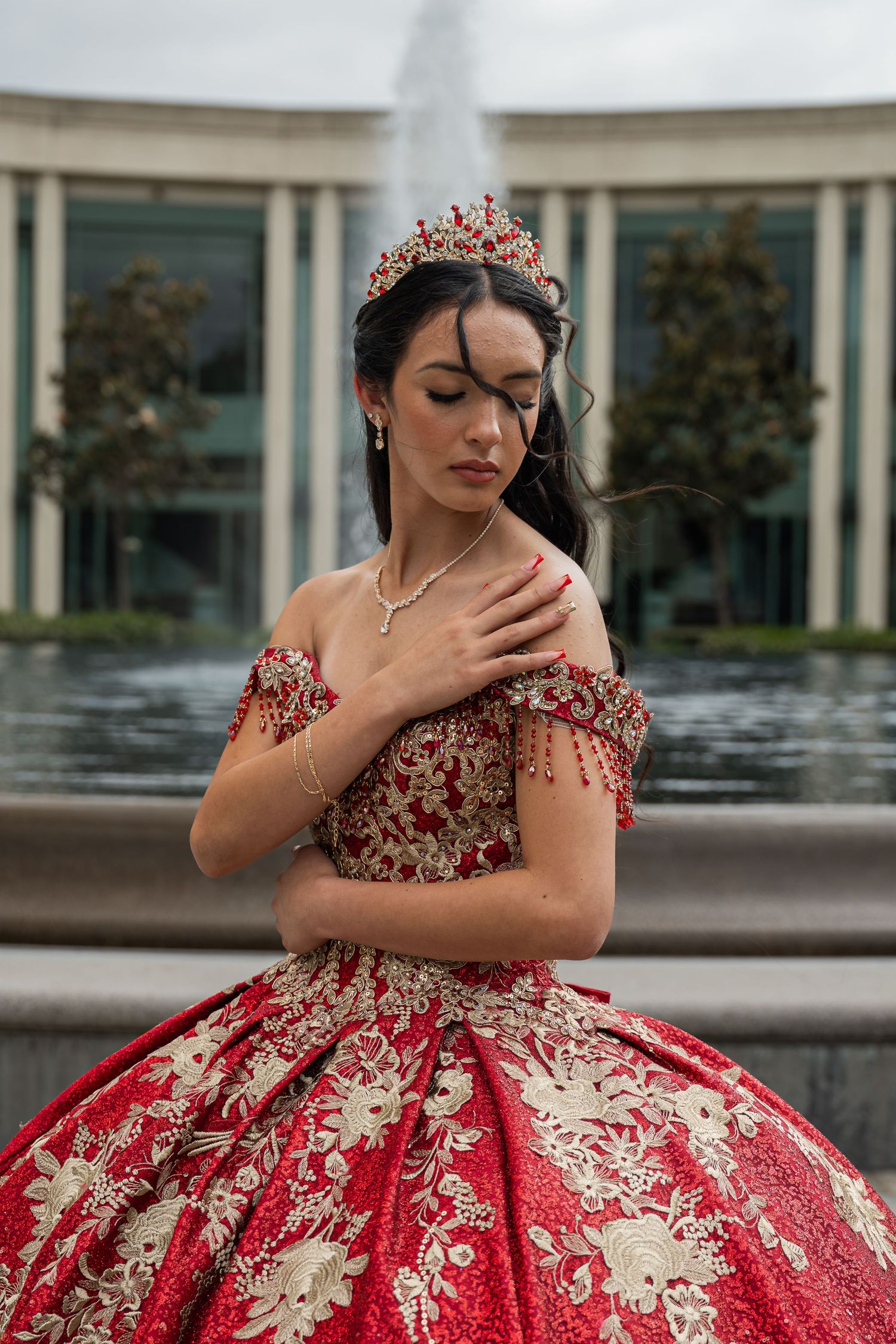 Young woman in red and gold quinceañera gown with crown poses by fountain.