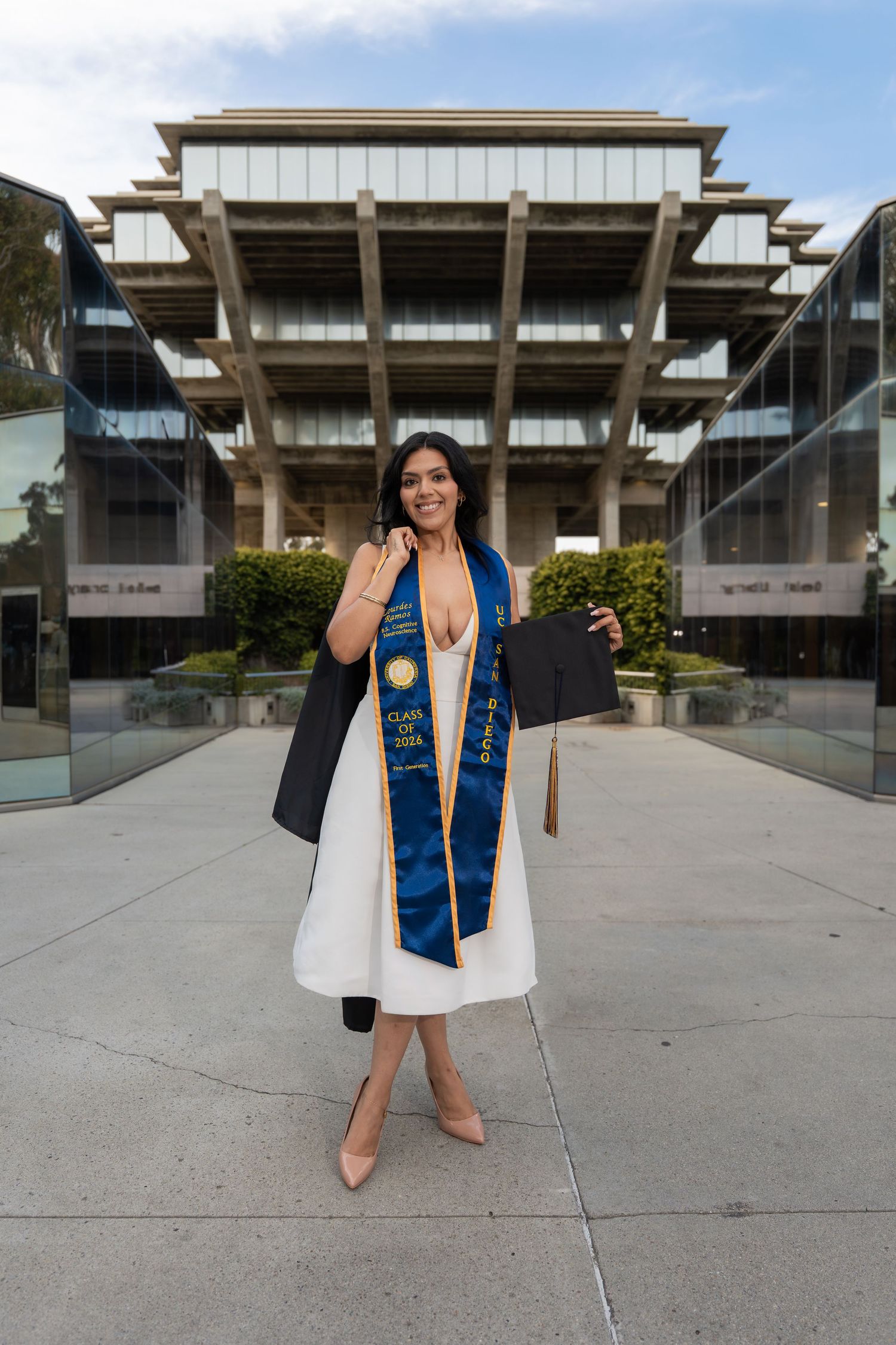 Smiling graduate in white dress and blue sash holds cap and gown outside Geisel Library at UC San Diego.