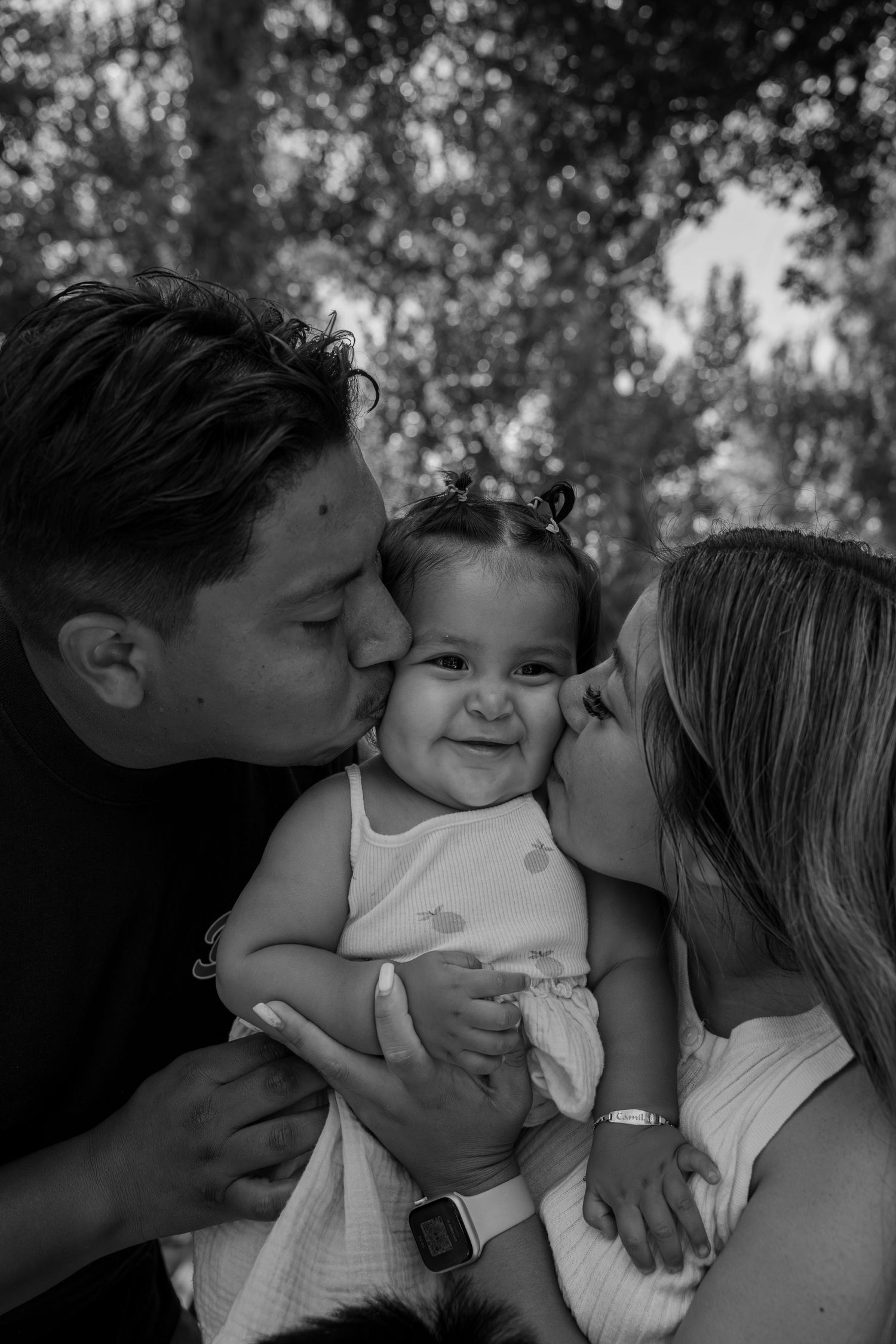 Black and white photo of parents kissing their smiling baby girl on both cheeks outdoors.