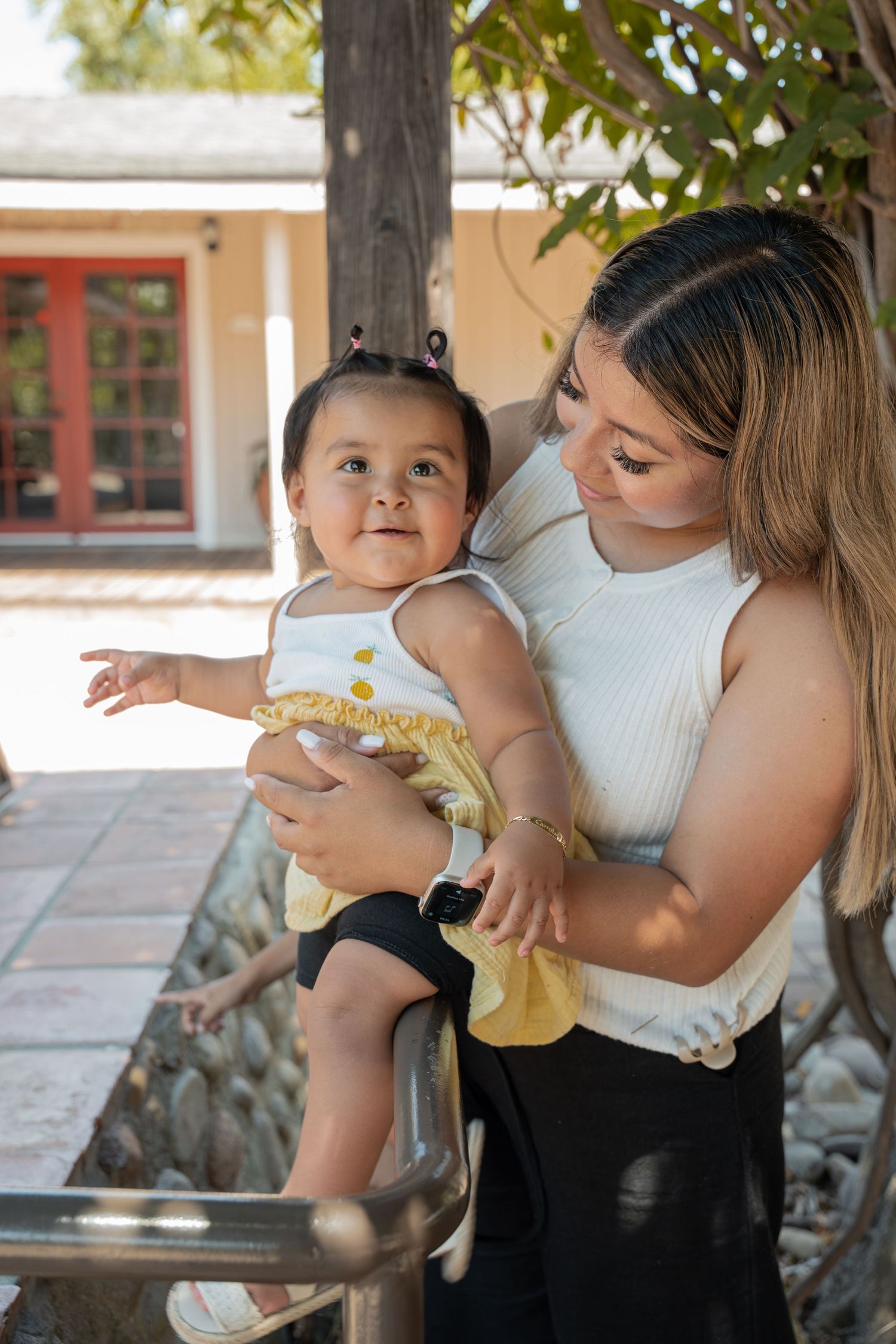 Young mother in white top holds her curious baby girl outdoors near a garden patio on a sunny day.