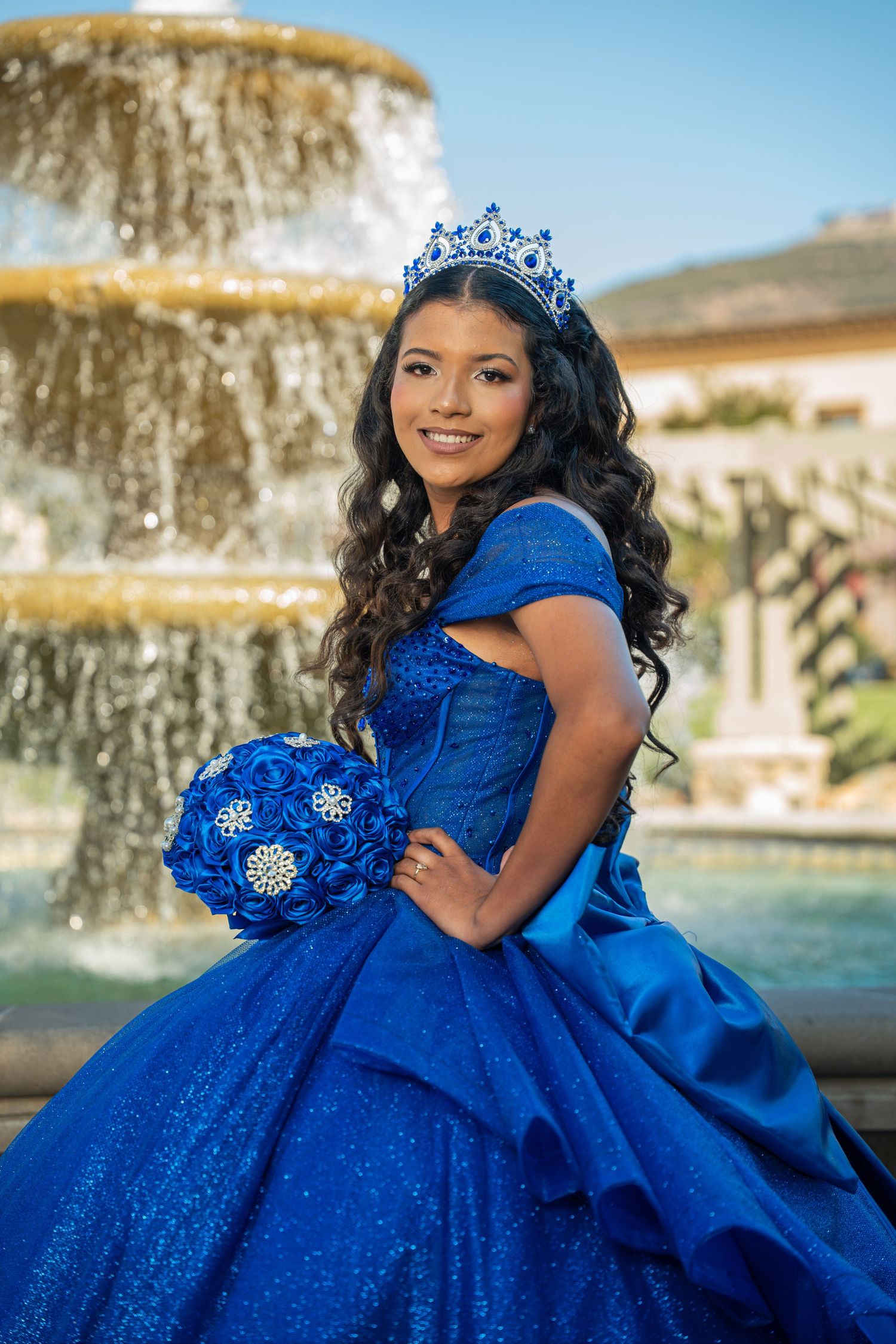Smiling quinceañera in a royal blue ball gown and tiara holds a bouquet in front of a fountain.