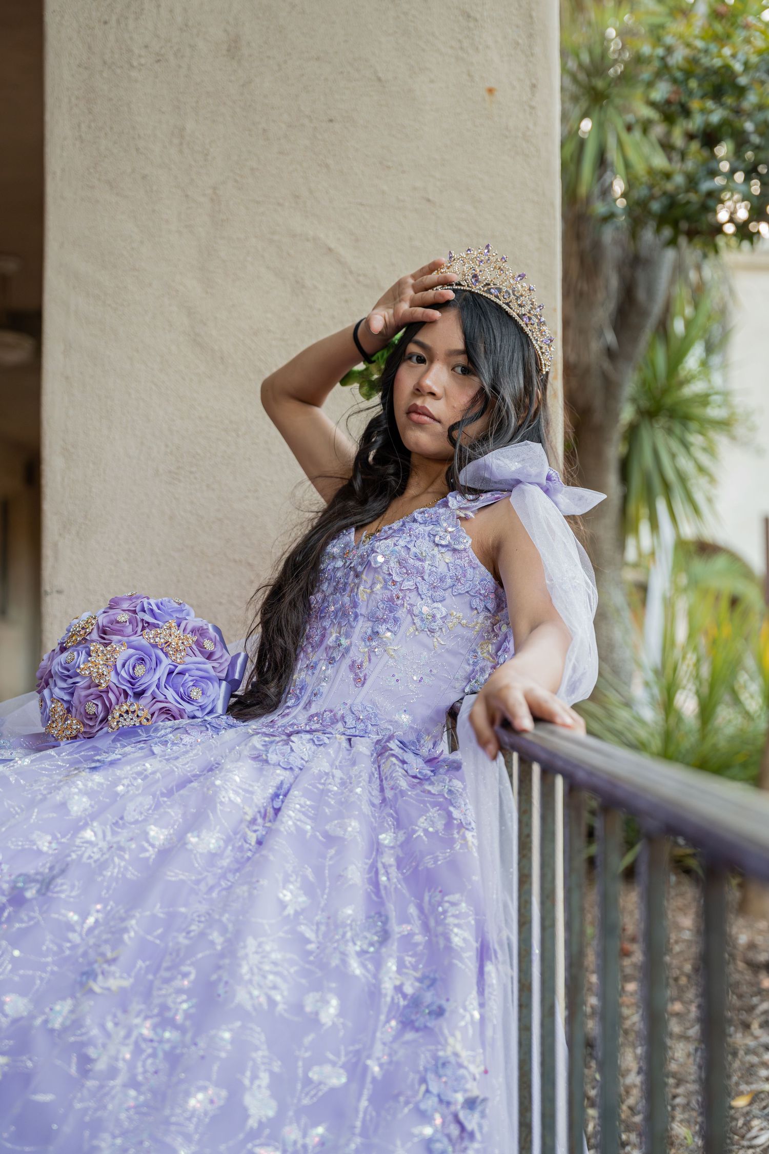 Teenage girl in lavender floral quinceañera gown and tiara crown poses on balcony with purple rose bouquet.