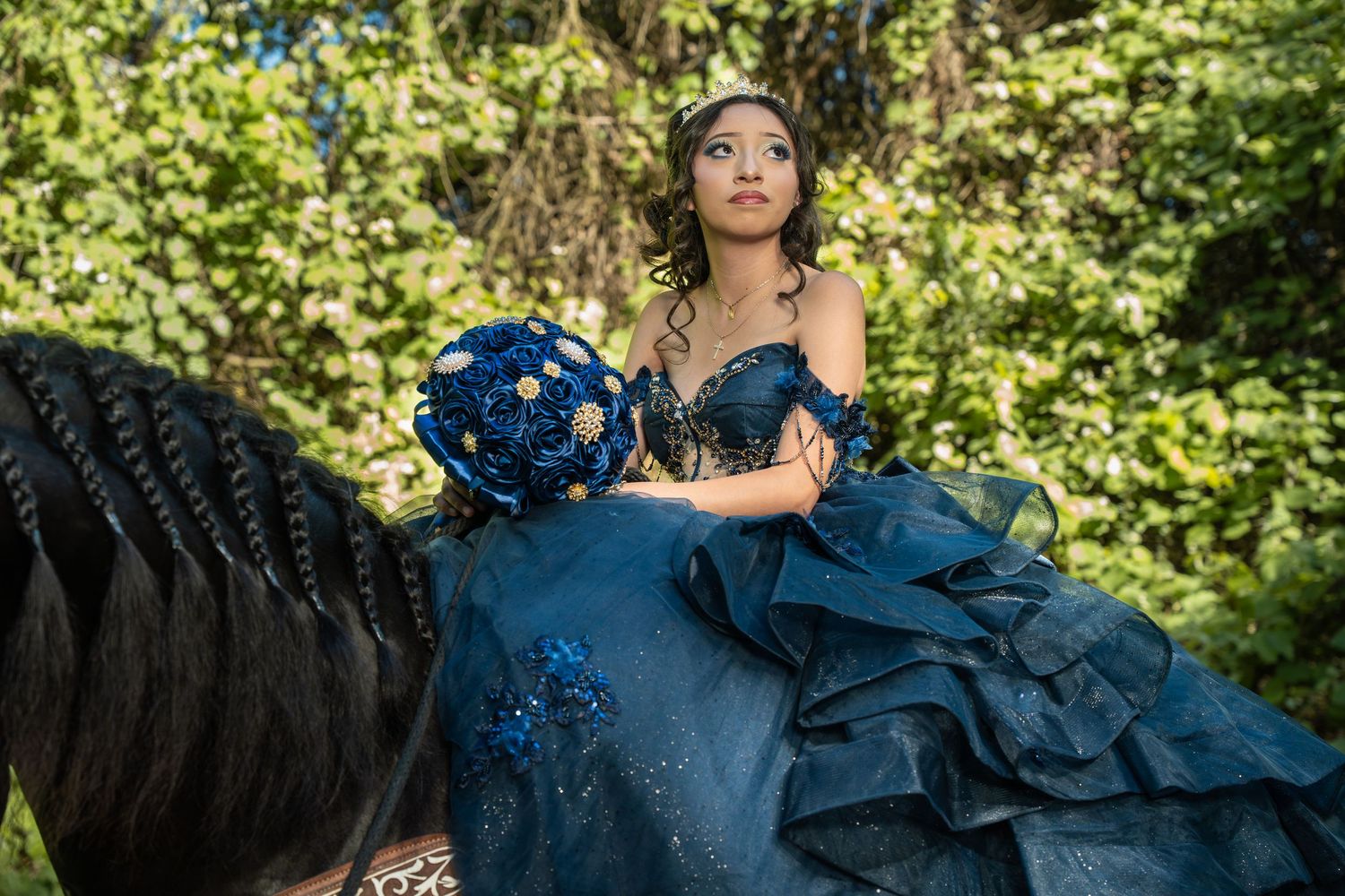 Quinceañera girl in dark blue ruffled gown holds blue bouquet while seated on horse surrounded by greenery.