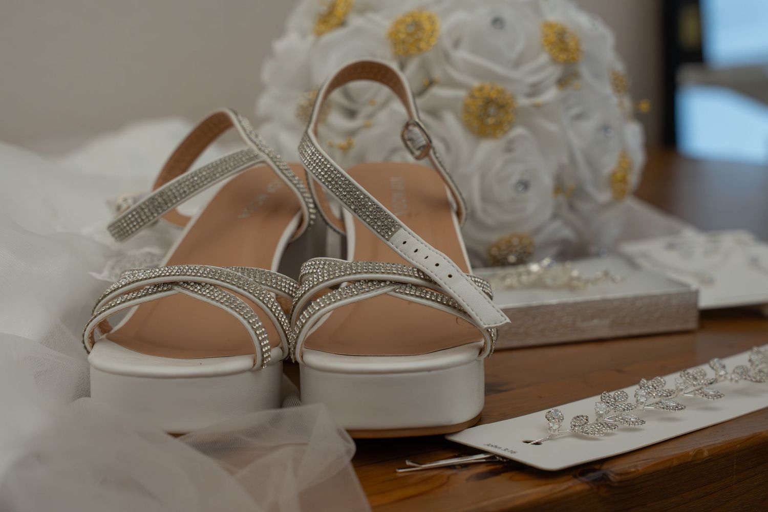 White rhinestone wedding sandals displayed on a wooden surface with a white floral bouquet and clutch in the background.