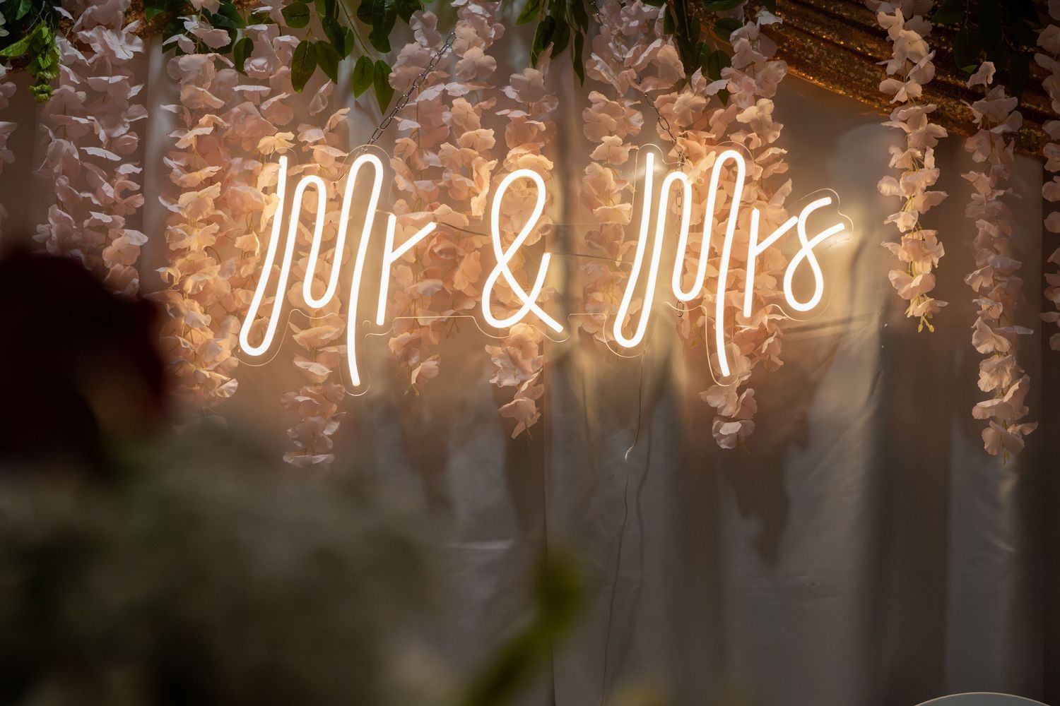 Glowing Mr and Mrs neon sign surrounded by hanging white wisteria flowers at romantic wedding venue.